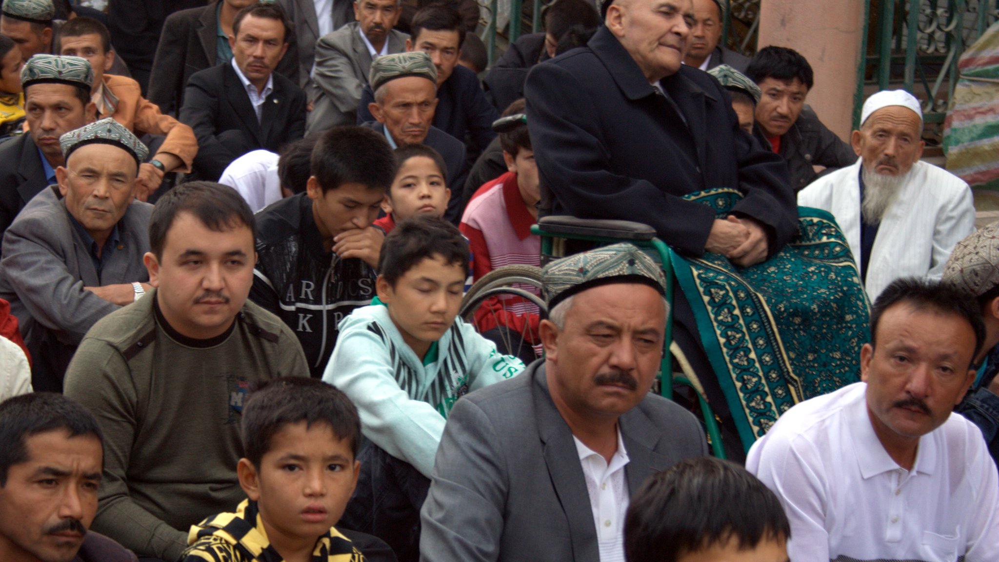 L'image montre un groupe de personnes assises, probablement lors d'un rassemblement ou d'une prière. On voit des hommes de différentes générations, certains portant des chapeaux traditionnels. Au centre, un homme âgé est assis sur une chaise, entouré de jeunes et d'adultes qui semblent attentifs. L'ambiance est sérieuse et respectueuse, reflétant un moment important pour la communauté.