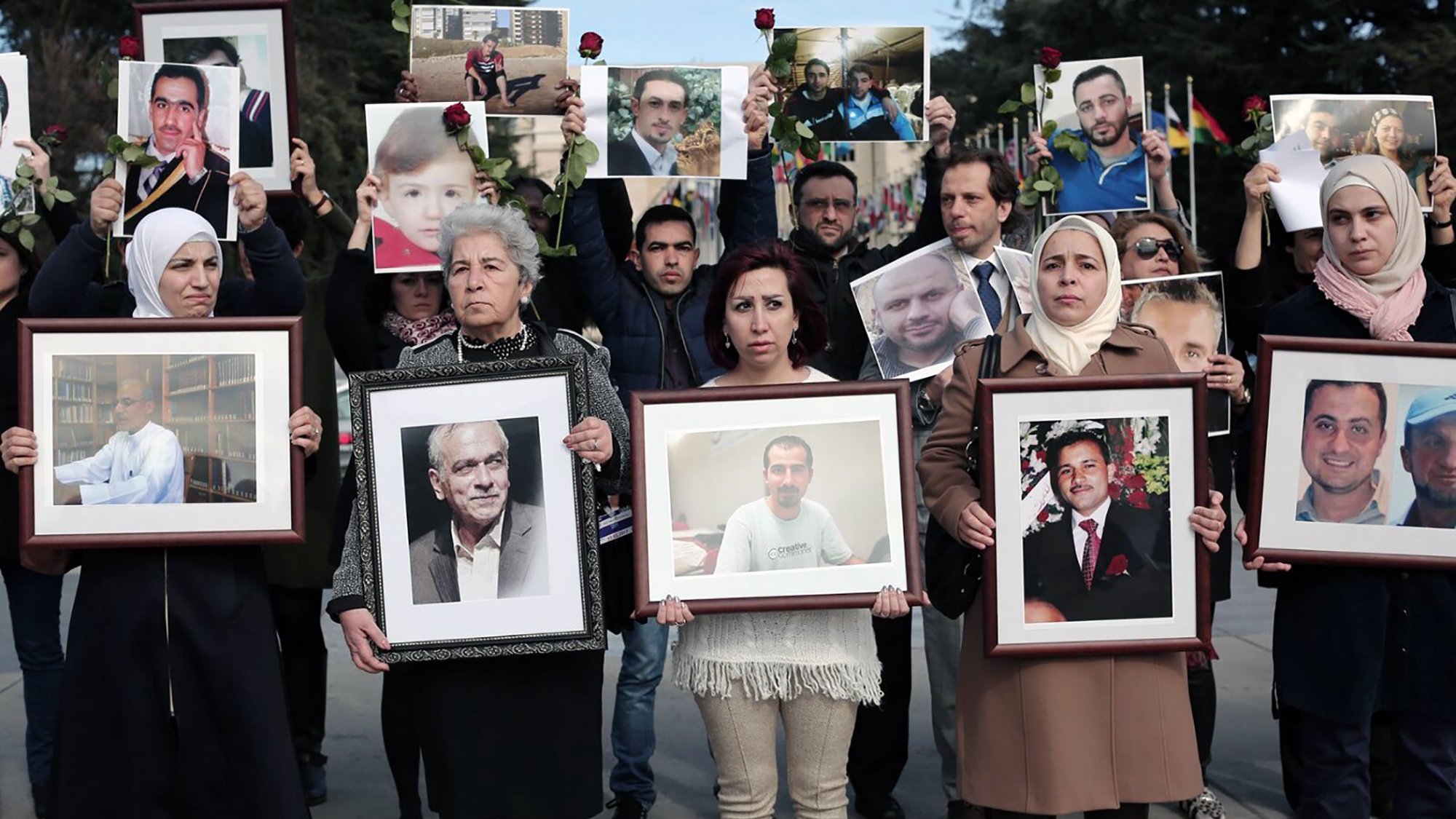 L'image montre un groupe de personnes participant à une manifestation ou un hommage. Elles tiennent des portraits de différents individus, certains avec des roses. Les expressions des participants semblent sérieuses et engagées, suggérant qu'ils rendent hommage à des personnes disparues ou aux victimes d'un conflit. Le décor en arrière-plan semble être en plein air, et plusieurs drapeaux sont visibles. Les vêtements des manifestants varient, avec quelques femmes portant des foulards.