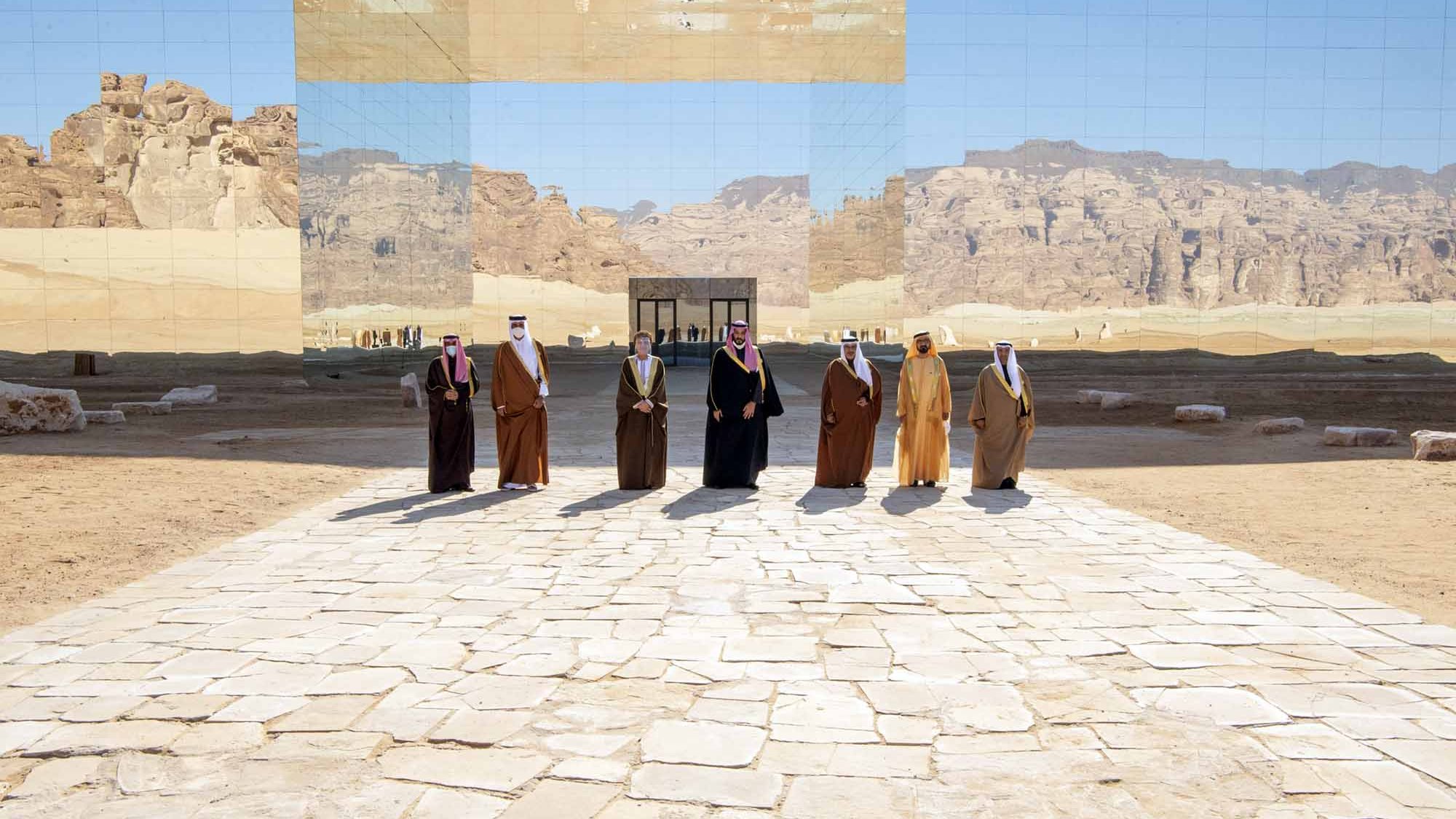 L'image montre un groupe d'individus en vêtements traditionnels, se tenant sur un chemin en pierre au milieu d'un paysage désertique. En arrière-plan, une grande structure réfléchissante fait un miroir du paysage environnant, créant un effet visuel intéressant. Les montagnes et le ciel sont également visibles, ajoutant une dimension naturelle à la scène. L'atmosphère semble solennelle et digne.