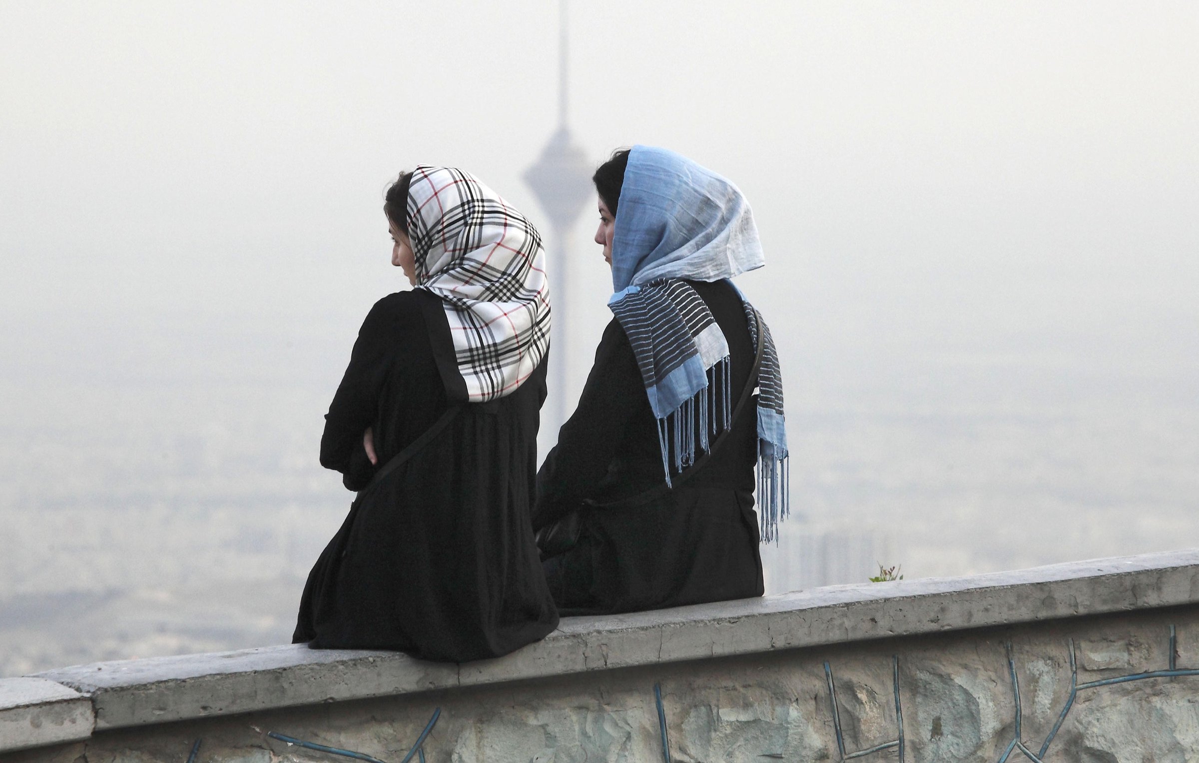 L'image montre deux femmes assises sur un mur, avec le dos tourné à la caméra. Elles portent des vêtements longs et des foulards, l'un d'eux étant à motifs. En arrière-plan, on aperçoit une ville embrumée avec ce qui semble être une tour, ajoutant un aspect urbain à la scène. L'atmosphère est paisible, avec une lumière douce, suggérant un moment de réflexion ou de contemplation.
