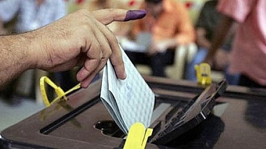 L'image montre une main avec un doigt marqué de violet, symbole d'un vote. La personne insère un bulletin de vote dans une urne électorale. En arrière-plan, on peut voir d'autres individus, probablement en train d'attendre ou de voter eux aussi. L'atmosphère semble sérieuse et engageante, illustrant un moment clé du processus démocratique.