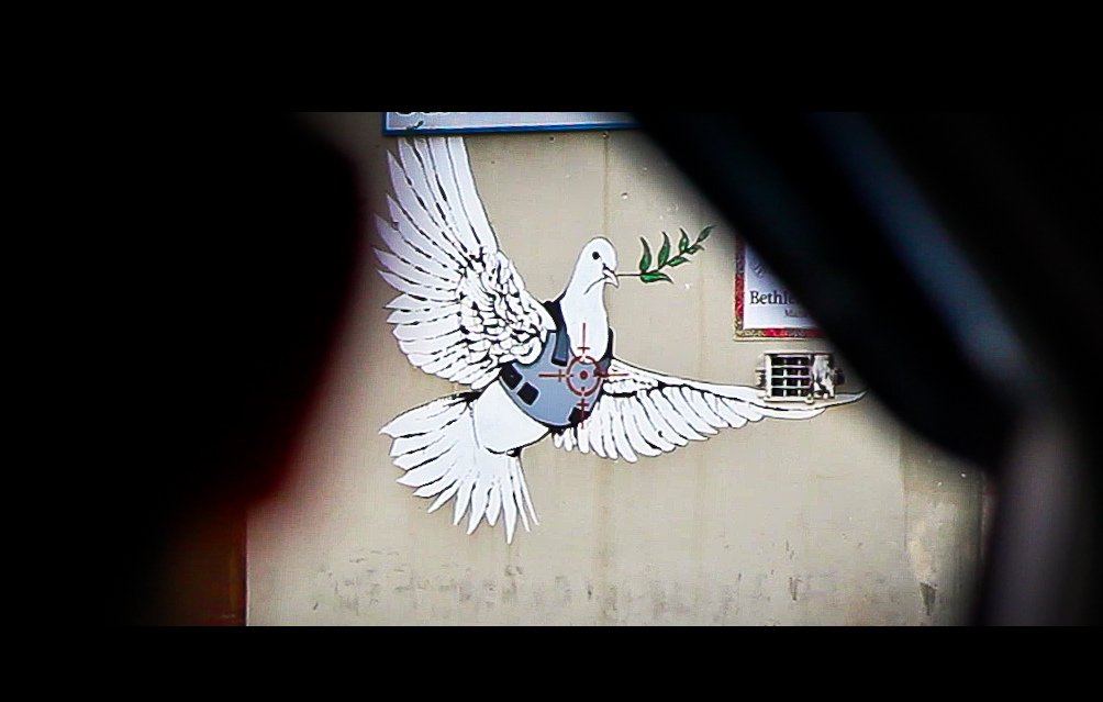 L'image montre une fresque murale représentant une colombe aux grandes ailes déployées. La colombe tient une branche d'olivier dans son bec, symbole de paix. Elle semble être stylisée avec des éléments modernes, comme un gilet pare-balles, ce qui pourrait faire allusion à des thèmes de conflits ou de sécurité. Le fond est neutre et sans éléments distracteurs, mettant en valeur la colombe et son message symbolique.