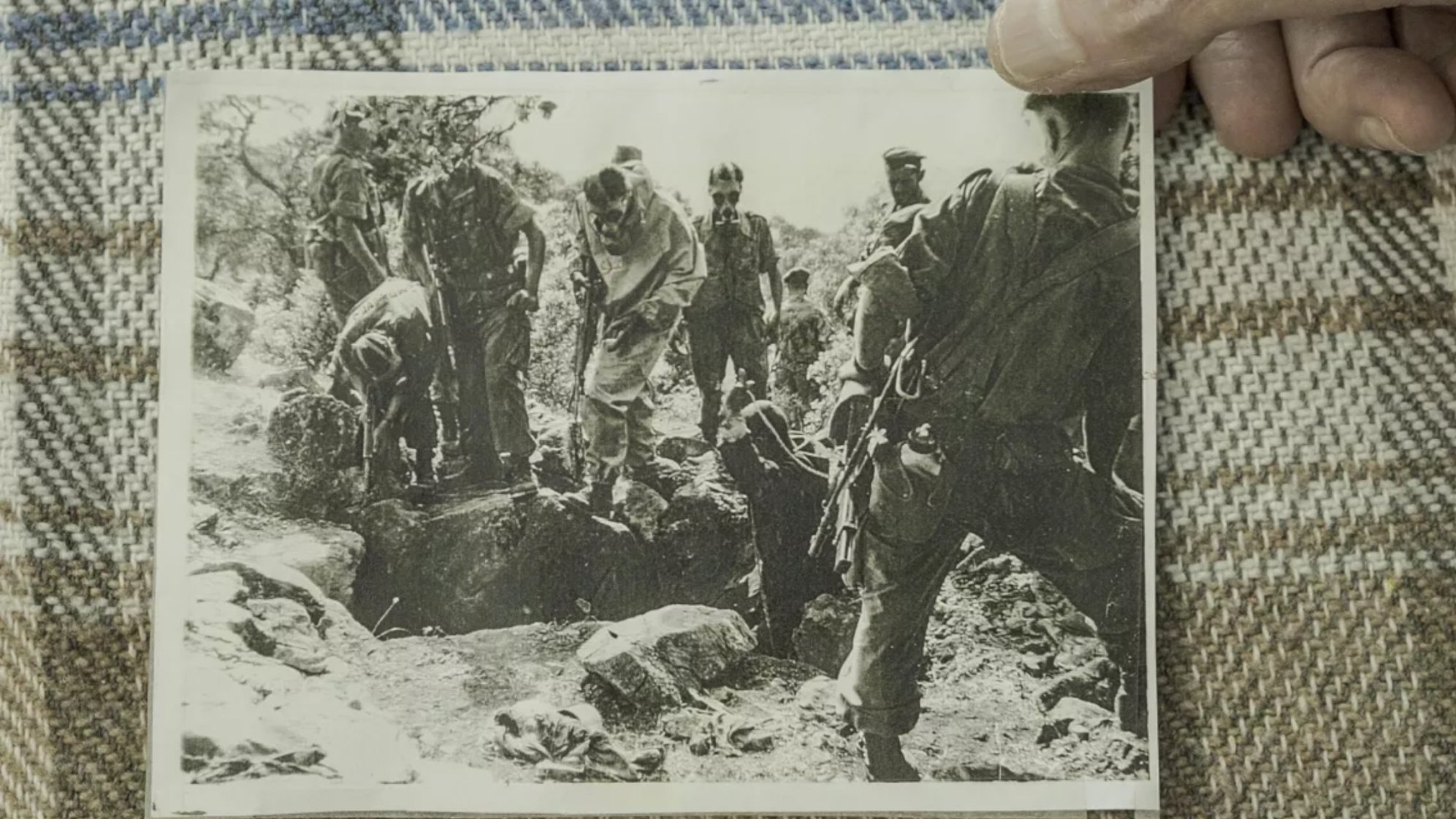 L'image montre un groupe de personnes en uniforme militaire, probablement des soldats, rassemblés autour d'un trou dans le sol. Certains semblent être en train de creuser ou d'examiner le terrain, tandis que d'autres observent. L'ambiance est sérieuse, indicative d'une situation d'urgence ou de conflit. Le tout est en noir et blanc.
