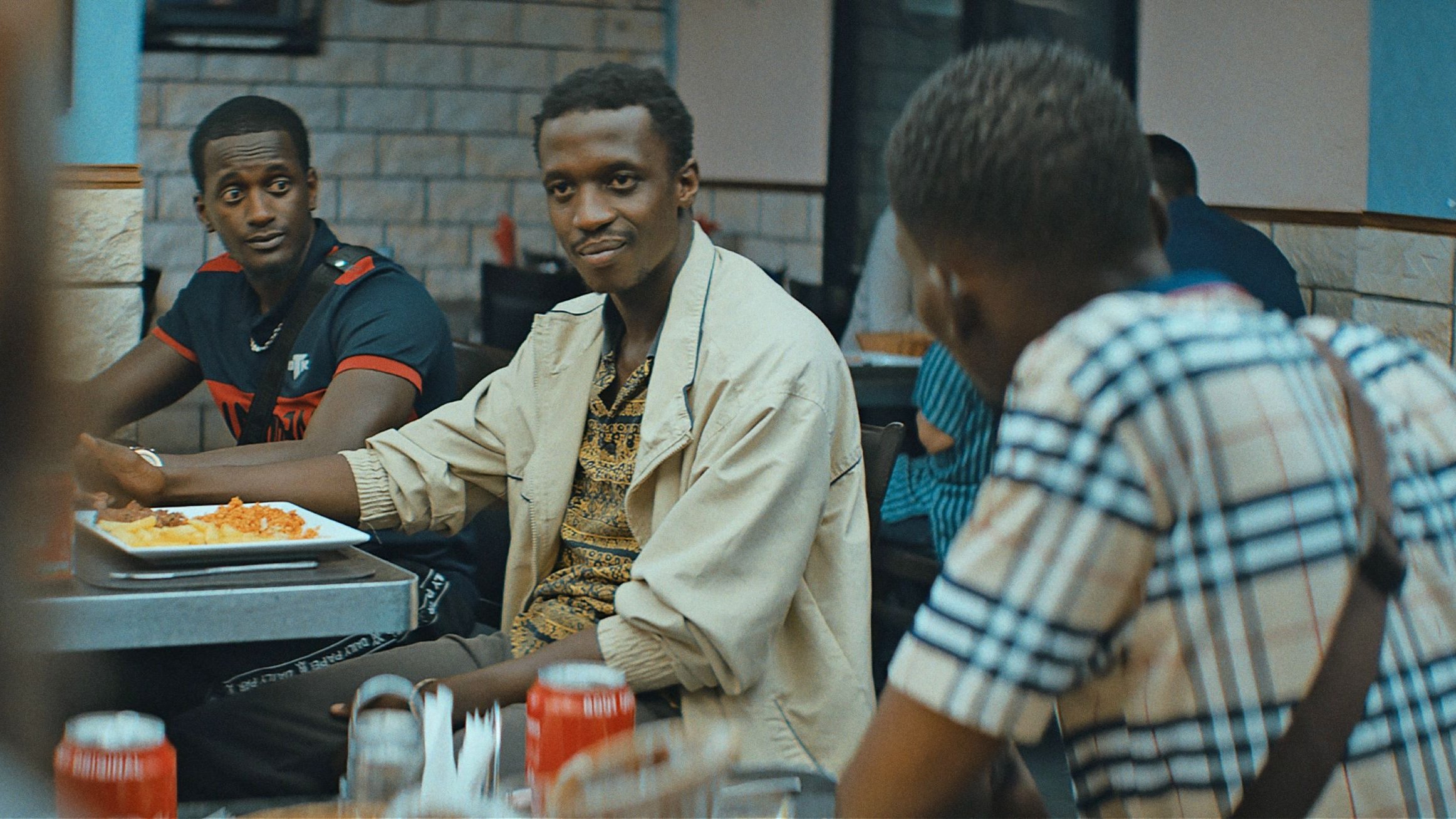 L'image montre un groupe de personnes assises dans un restaurant. Au premier plan, un homme souriant, portant une chemise claire et un motif coloré, semble engager une conversation. Deux autres hommes sont également visibles : l'un avec un maillot noir et rouge, et l'autre portant une chemise à carreaux. Les tables sont garnies de plats de nourriture, et l'ambiance est détendue, typique d'un moment convivial entre amis.