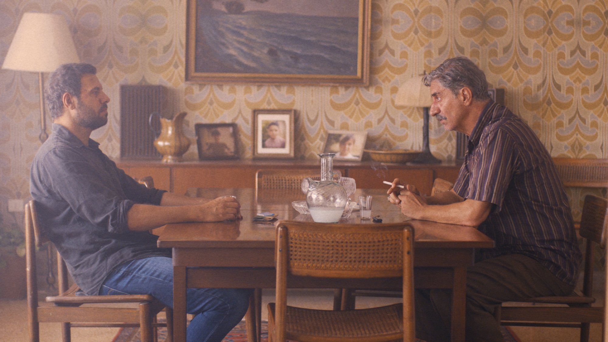 L'image montre deux hommes assis à une table en bois dans une pièce décorée avec un papier peint aux motifs rétro. L'ambiance est intérieure et intimiste, avec une lampe à côté et des photos accrochées au mur. Les deux hommes semblent engagés dans une conversation sérieuse. Le premier homme, à gauche, est vêtu d'une chemise sombre et regarde l'autre. Le deuxième homme, à droite, fume une cigarette et semble pensif. L'éclairage doux ajoute à l'atmosphère.