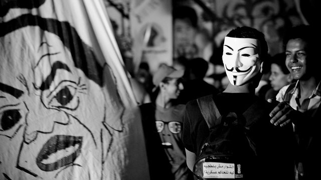 L'image montre une scène de rassemblement ou de manifestation. Un groupe de personnes est réuni, et au premier plan, une personne porte un masque stylisé, souvent associé au mouvement Anonymous. En arrière-plan, on peut voir une grande bannière avec des illustrations ou des visages, probablement liés à un message politique. L'atmosphère semble dynamique, avec des expressions de joie ou de solidarité parmi les participants. Le tout est en noir et blanc, ce qui accentue l'intensité de la scène.