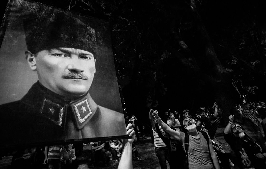 L'image montre un rassemblement nocturne où des gens tiennent un grand portrait d'une figure historique. Les participants semblent exprimer leur admiration ou leur respect, levant le poing en signe de solidarité ou de protestation. L'atmosphère est festive mais aussi solennelle, avec des ombres et une luminosité faible qui accentue l'intensité du moment.