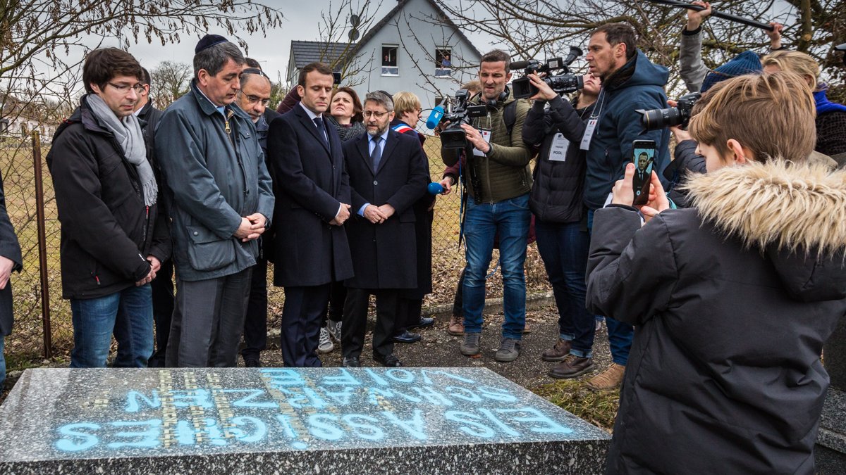 L'image montre un groupe de personnes rassemblées autour d'une stèle ou d'une tombe. Parmi elles, on peut distinguer des individus en tenue formelle, semblant assister à une cérémonie hommage. Un enfant tient un téléphone pour filmer ou prendre une photo. Des journalistes, avec des caméras et des micros, sont également présents, suggérant que l'événement est médiatisé. En arrière-plan, on aperçoit une maison et des arbres, indiquant un cadre extérieur.