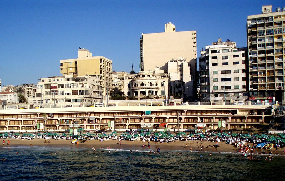 L'image montre une plage animée avec de nombreux baigneurs profitant du soleil et de la mer. On peut voir des parasols disposés sur le sable, ainsi que des hôtels et des immeubles en arrière-plan. L'architecture des bâtiments est variée, avec des structures anciennes côtoyant des constructions plus modernes. Le ciel est dégagé, et l'atmosphère est vivante, typique d'une destination balnéaire populaire.