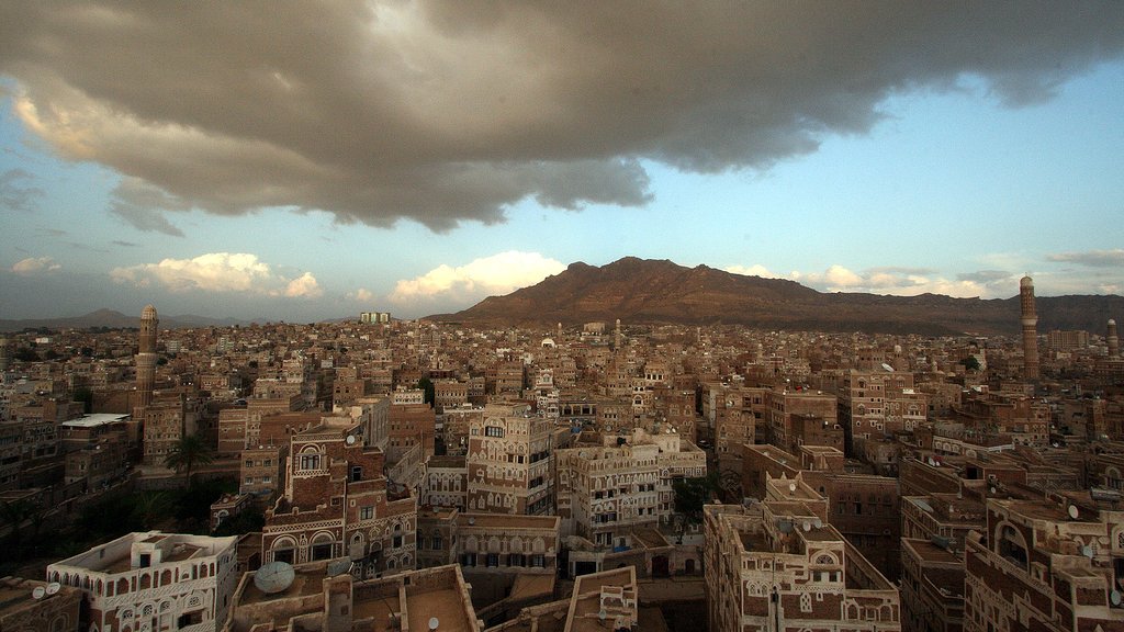 L'image montre une vue panoramique d'une ville traditionnelle, probablement située au Yémen, avec des bâtiments en adobe et des structures architecturales distinctives. On aperçoit des montagnes en arrière-plan, sous un ciel nuageux qui laisse entrevoir quelques éclaircies. La lumière du soleil couchant semble illuminer une partie de la ville, mettant en valeur les détails des constructions. L'ensemble dégage une atmosphère calme et pittoresque, reflétant la beauté du paysage urbain.