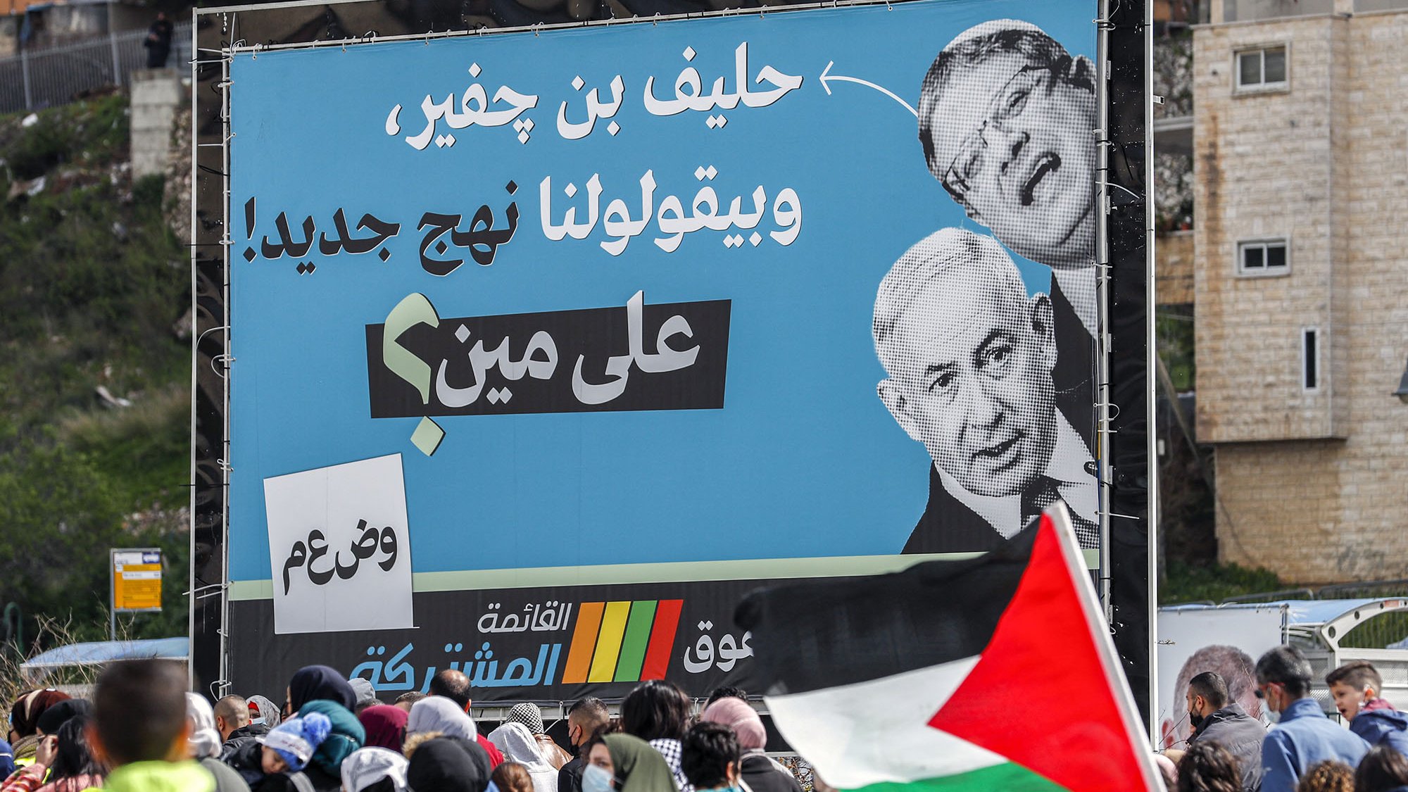L'image montre une grande affiche sur un panneau publicitaire, présentant des portraits de deux hommes, dont l'un semble être un homme politique conversationnel. En arrière-plan, on peut voir une foule de personnes, certaines tenant des drapeaux palestiniens. L'affiche semble véhiculer un message politique, avec des slogans en arabe, suggérant un contexte de mobilisation ou de protestation. L'ambiance est dynamique, reflétant une situation de rassemblement public.