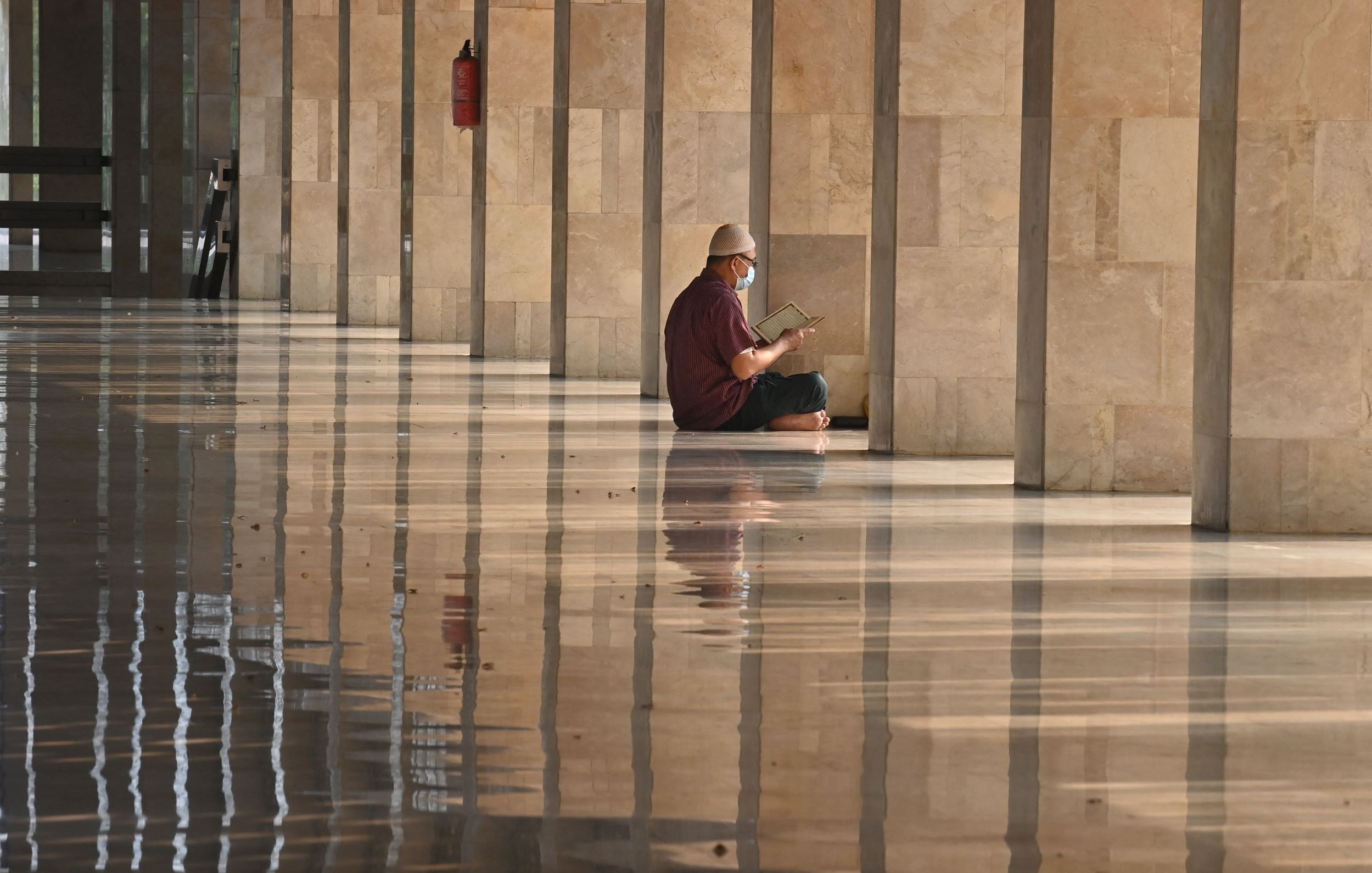 L'image montre un homme assis sur le sol d'un espace intérieur, probablement un grand hall ou une mosquée, avec des murs de pierre ainsi qu'un sol brillant et réfléchissant. L'homme porte un chapeau et lit un livre, et l'environnement est calme et serein. Il semble être plongé dans sa lecture, tandis que la lumière naturelle crée des reflets sur le sol.