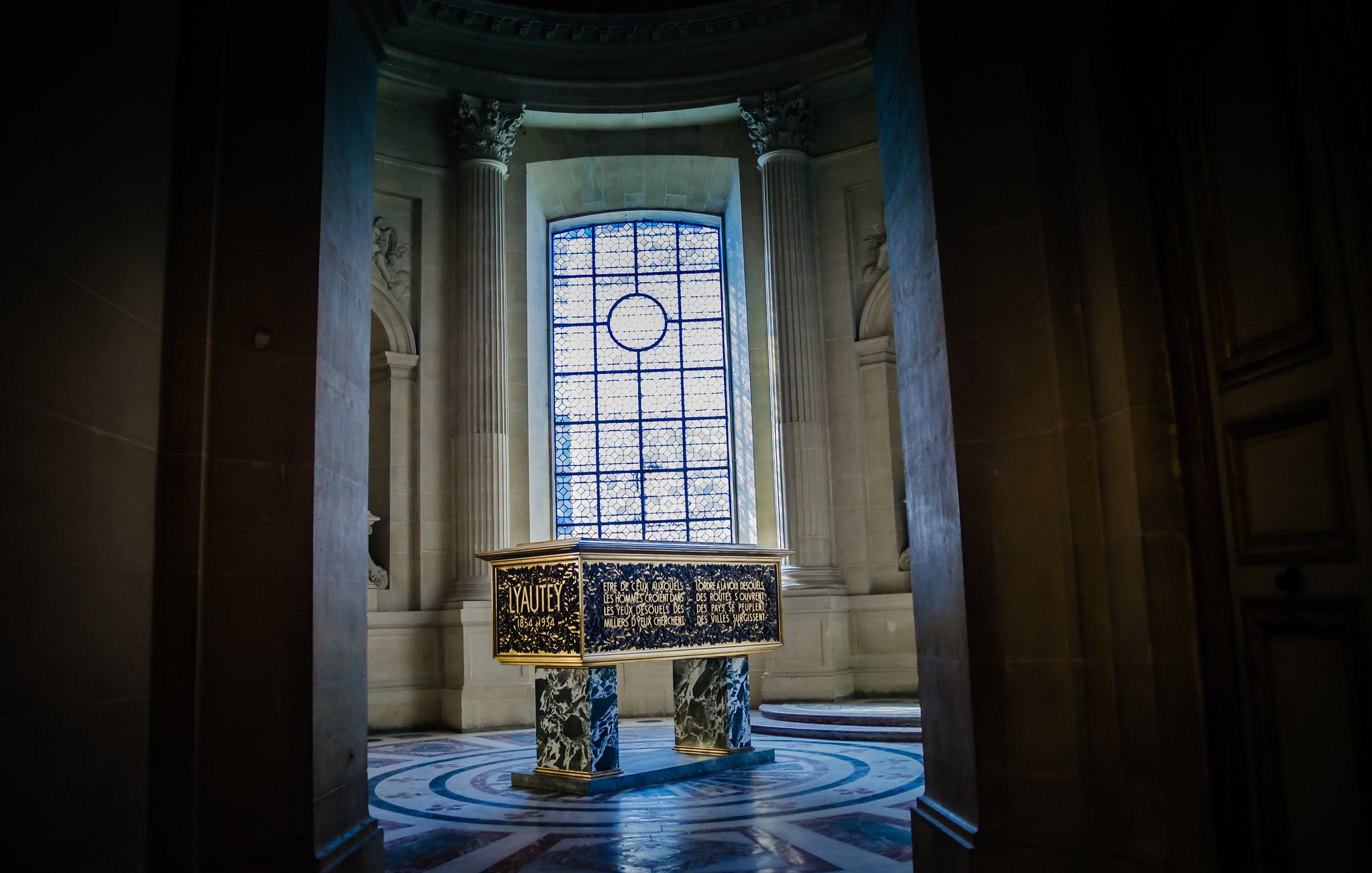L'image montre une pièce intérieure d'un bâtiment majestueux, probablement un palais ou un musée. Au centre, il y a un grand meuble ou une caisse ornée de motifs décoratifs dorés et peints. Les murs sont en pierre, avec de très grandes fenêtres, à travers lesquelles entre une lumière douce, créant une atmosphère solennelle. Les colonnes et le sol richement décoré ajoutent à la grandeur de l'espace.