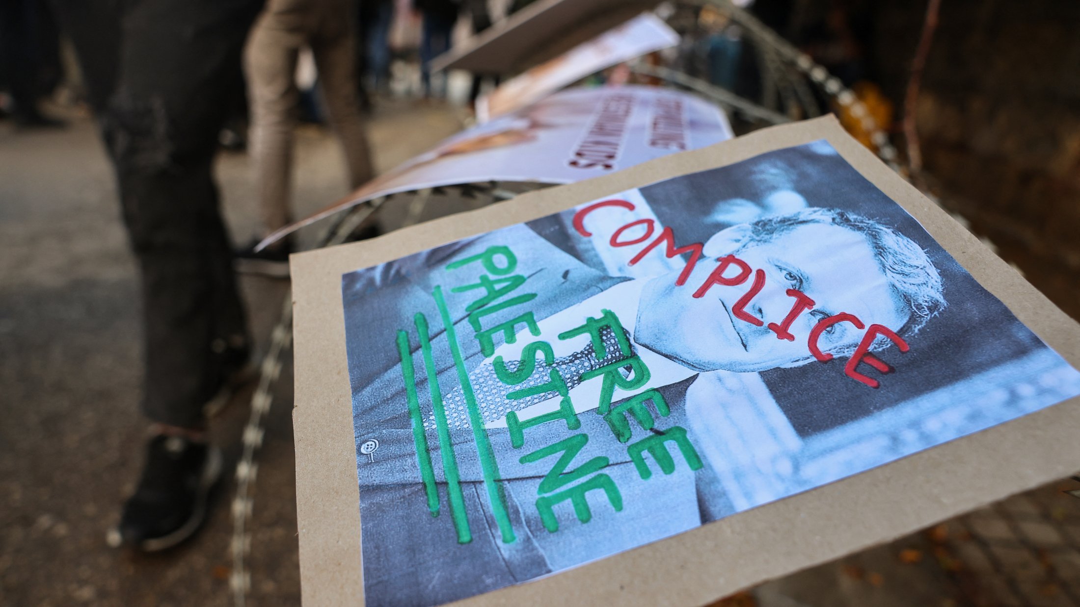 L'image montre des affiches placées sur une barrière, probablement dans le cadre d'une manifestation. L'une des affiches présente un portrait en noir et blanc avec les mots "COMPLICE" et "FREE PALESTINE" écrits en rouge et vert. L'environnement semble être celui d'une manifestation ou d'un rassemblement, avec plusieurs personnes en arrière-plan.