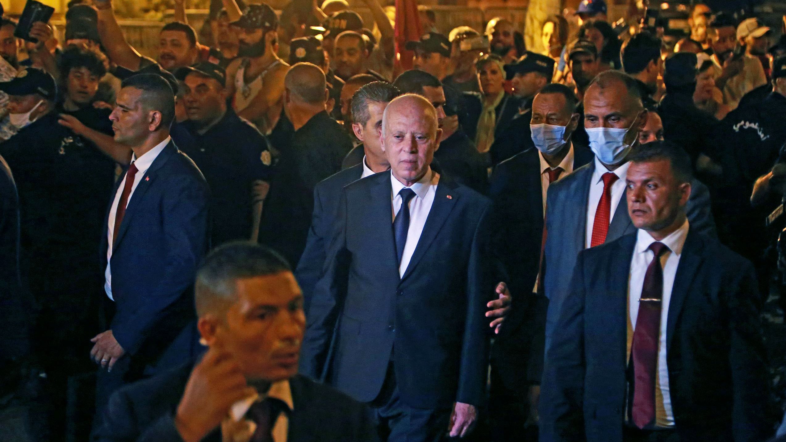 L'image montre un groupe de personnes rassemblées autour d'un homme en costume noir, qui semble être une figure politique ou publique. Il est entouré de plusieurs membres de ce qui semble être une équipe de sécurité, tous vêtus de costumes sombres. Les gens en arrière-plan semblent attentifs et certains utilisent des téléphones pour prendre des photos. L'ambiance paraît animée, avec un éclairage qui suggère qu'il fait sombre.