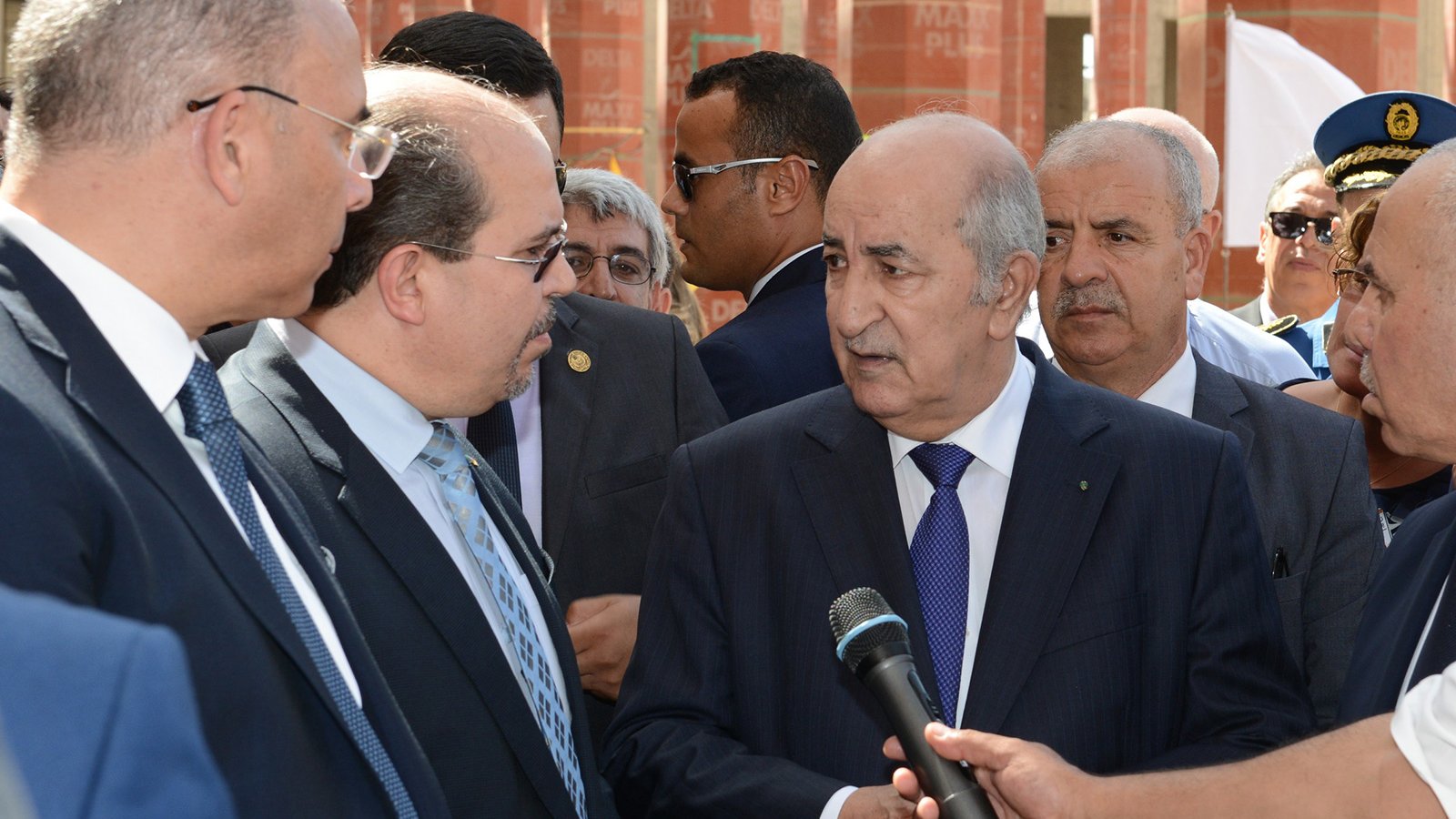 L'image montre un groupe d'hommes en costume, probablement en train de discuter. Au centre, l'un des hommes semble occuper une position de leader, tandis que les autres l'écoutent attentivement. Ils semblent être dans un cadre formel, peut-être lors d'une visite officielle ou d'un événement important. L'environnement en arrière-plan suggère un lieu institutionnel, avec des structures architecturales visibles.