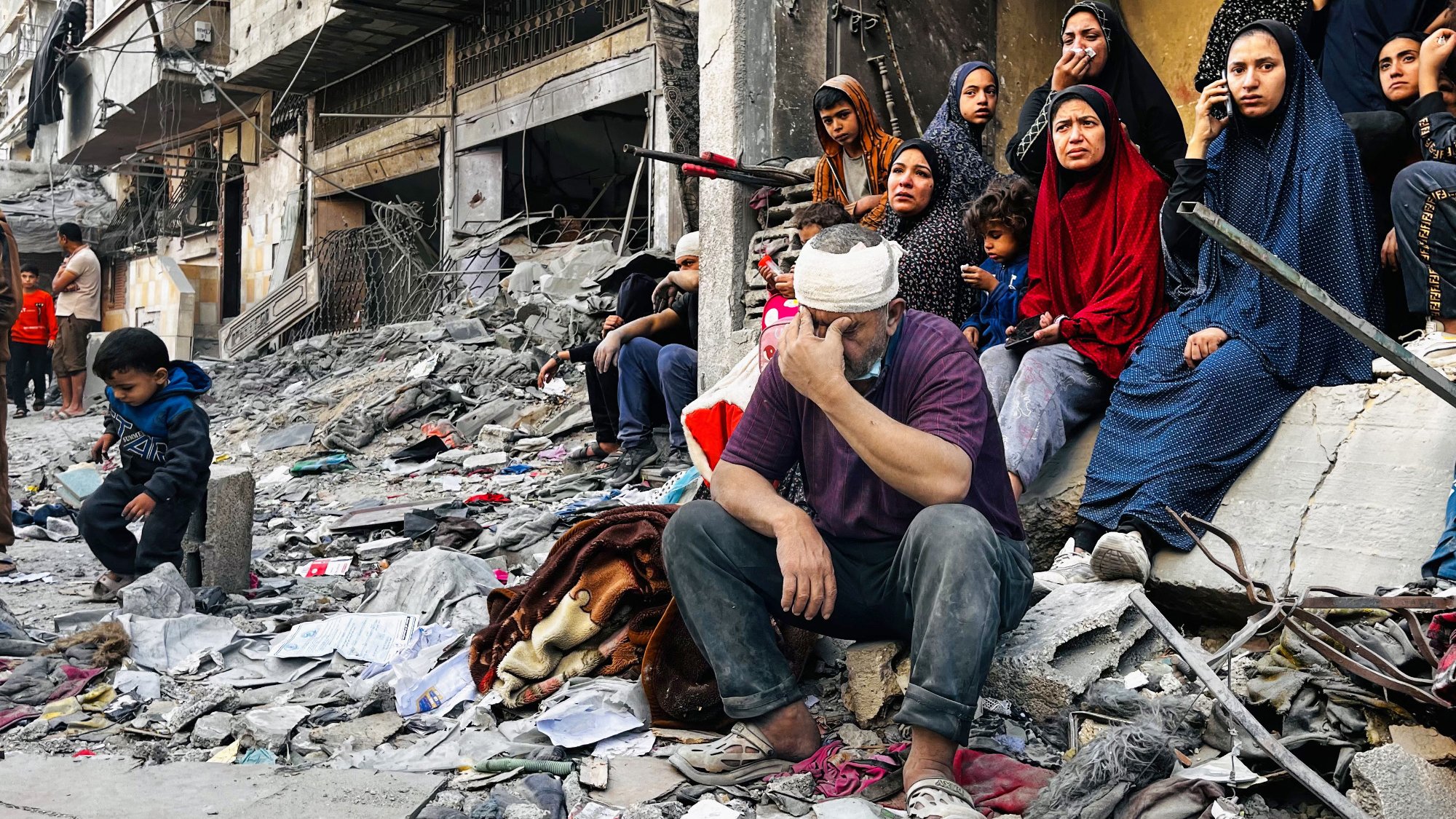 L'image montre un groupe de personnes assises sur des décombres dans une zone de conflit. Au premier plan, un homme, la tête bandée, exprime une grande tristesse ou du désespoir, tandis qu'à proximité, plusieurs femmes en vêtements sombres regardent vers l'objectif, certaines avec une expression préoccupée. Un petit garçon est assis un peu plus loin, semblant perdu dans ses pensées. L'environnement est chaotique, avec des ruines et des débris autour, suggérant des dommages importants causés par des événements récents. L'atmosphère générale est lourde, reflétant la douleur et la résilience des personnes présentes.