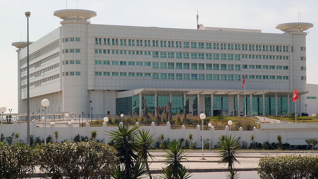 L'image montre un bâtiment moderne et imposant, probablement un centre administratif ou gouvernemental. Il est de couleur blanche avec de grandes fenêtres rectangulaires, donnant une impression de fonctionnalité et de sobriété. Le design architectural est contemporain, avec des formes géométriques claires. À l'avant, il y a un espace ouvert avec des éléments paysagers tels que des plantes et des lampadaires. On peut également voir un drapeau, ce qui pourrait indiquer qu'il s'agit d'un bâtiment officiel. L'environnement semble propre et bien entretenu.