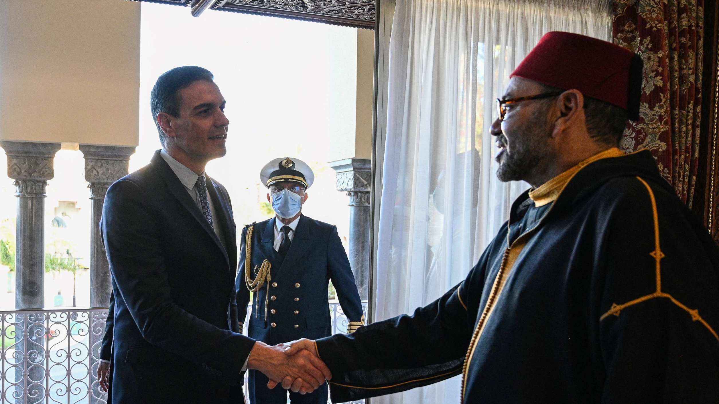 L'image montre deux hommes en train de se serrer la main dans un cadre intérieur élégant. L'un des hommes porte un costume, tandis que l'autre est habillé d'un habit traditionnel noir avec un chapeau rouge. En arrière-plan, un homme en uniforme militaire observe la scène. La lumière naturelle entre par une grande fenêtre avec des rideaux blancs, ajoutant une ambiance chaleureuse à la rencontre.