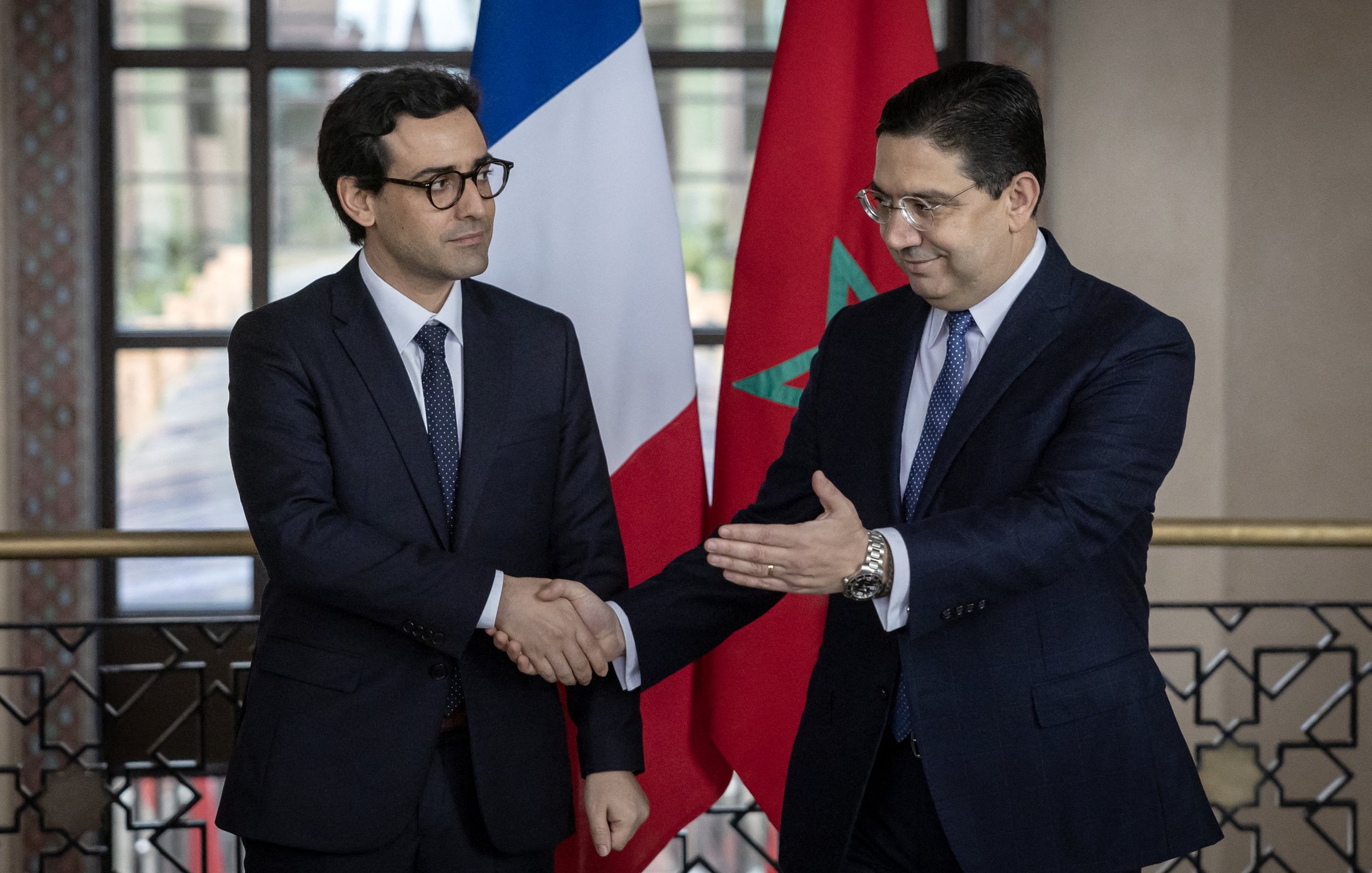 L'image montre deux hommes en costume, se serrant la main dans un cadre officiel. À l'arrière-plan, on peut voir un drapeau français à gauche et un drapeau marocain à droite. L'atmosphère semble être celle d'une rencontre diplomatique ou d'une conférence de presse, soulignant une interaction entre les deux pays.
