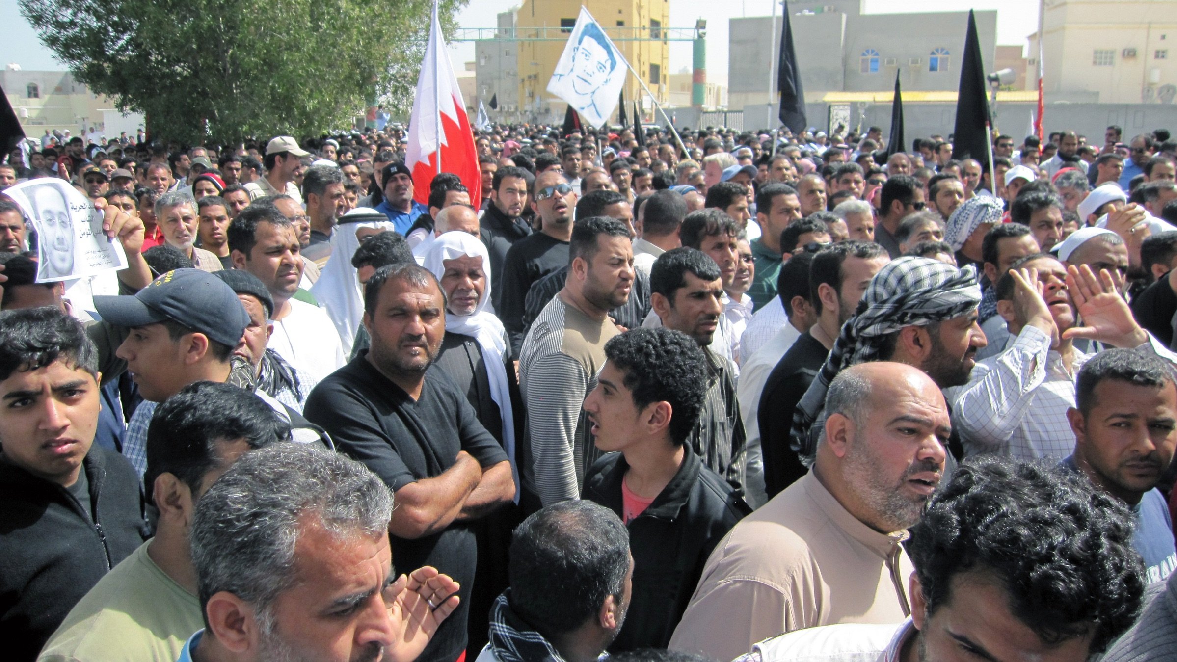 L'image montre une grande foule de personnes rassemblées, probablement lors d'une manifestation. Les participants semblent très engagés, certains tenant des drapeaux, y compris celui de Bahreïn. Les visages expriment diverses émotions, allant de la détermination à l'interrogation. L'environnement montre des bâtiments en arrière-plan, suggérant une zone urbaine. L'atmosphère générale dénote une forte implication politique ou sociale.