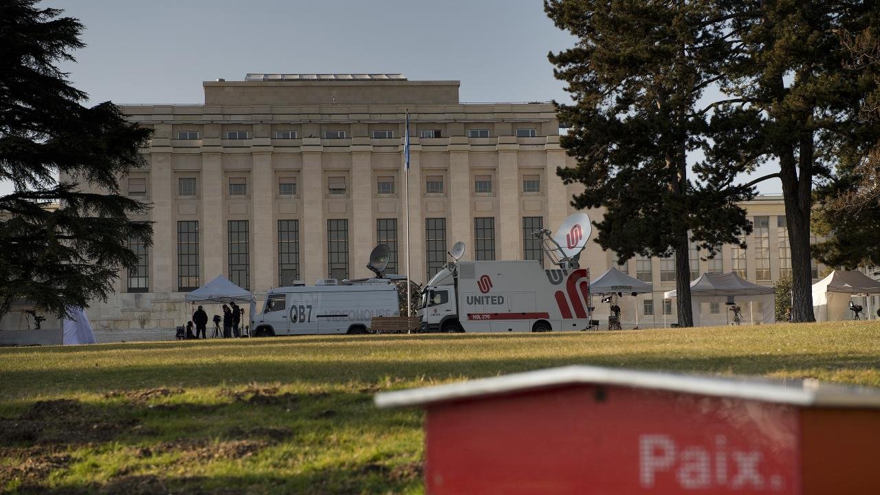 L'image montre un bâtiment imposant, probablement un palais ou un bâtiment gouvernemental, situé à Genève. À l'avant-plan, on aperçoit une petite structure rouge portant le mot "Paix". En arrière-plan, on distingue des véhicules de télévision et des tentes, indiquant qu'un événement ou une conférence pourrait avoir lieu. Le cadre est agrémenté d'arbres, suggérant un espace extérieur agréable.
