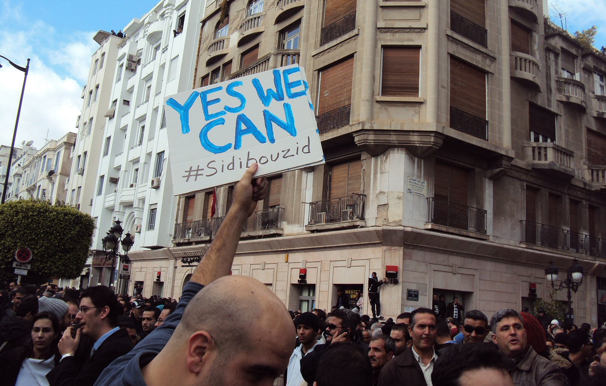 L'image montre une foule rassemblée dans une ville, probablement lors d'une manifestation. Au centre, une personne tient une pancarte avec le message "YES WE CAN" et le hashtag "#Sidibouzid". Les bâtiments en arrière-plan suggèrent un environnement urbain. L'atmosphère semble être celle d'un mouvement populaire, avec des participants engagés et déterminés.