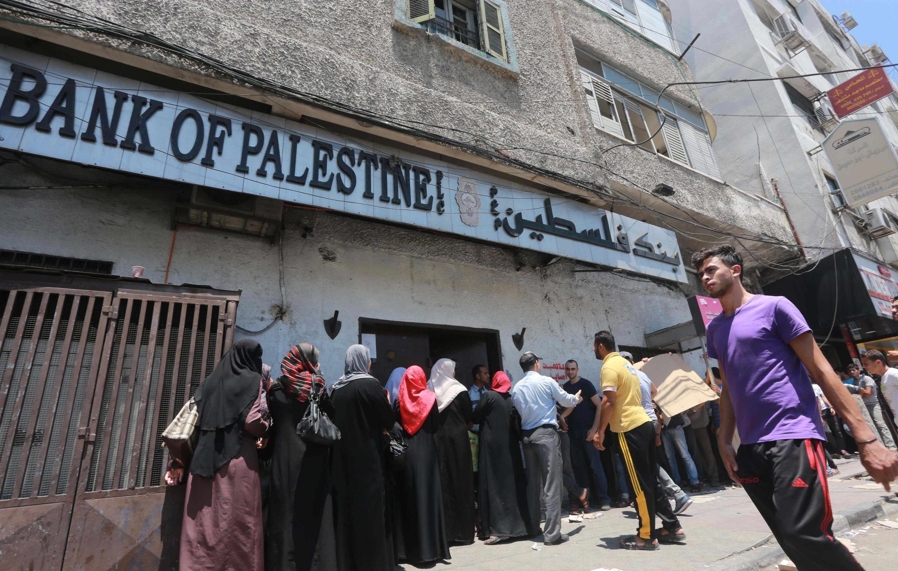 L'image montre une scène extérieure près d'un bâtiment portant l'inscription "Bank of Palestine". On y voit une file de personnes, principalement des femmes portant des vêtements traditionnels, attendant à l'extérieur de la banque. Certains hommes sont également présents, et on aperçoit un groupe d'individus qui se tiennent plus près de l'entrée. En arrière-plan, le bâtiment est ancien et les conditions semblent assez chaotiques, avec des câbles électriques visibles et des fenêtres en état d'usage. Le temps semble ensoleillé, suggérant une ambiance chaude.