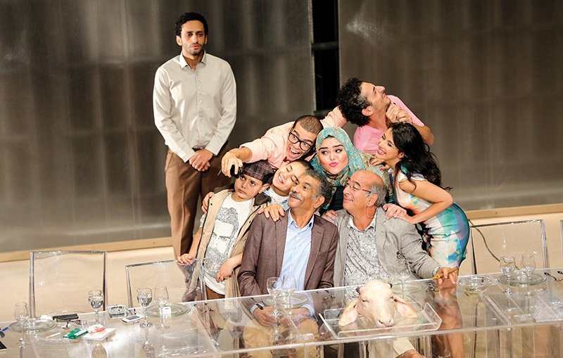 L'image montre un groupe de personnes rassemblées autour d'une table, apparemment lors d'un événement convivial. Il y a des adultes et des enfants, certains prenant la pose pour un selfie. Les expressions sont joyeuses et décontractées, avec des sourires sur presque tous les visages. À l'arrière-plan, un homme se tient debout, regardant la scène. La table est décorée avec des verres et un plat, ajoutant une ambiance festive à la scène. L'ensemble dégage une atmosphère d'amitié et de bonheur partagé.