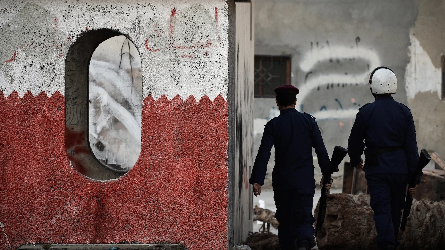 L'image montre deux policiers marchant le long d'un mur. Ce mur présente une décoration en rouge et blanc, avec une forme ovale découpée. En arrière-plan, plusieurs murs sont couverts de graffitis et apparaissent en mauvais état, témoignant d'une ambiance de tension ou de désolation. Les policiers, munis de casques et d'armement, semblent patrouiller dans une zone qui pourrait être marquée par des troubles ou des désordres.
