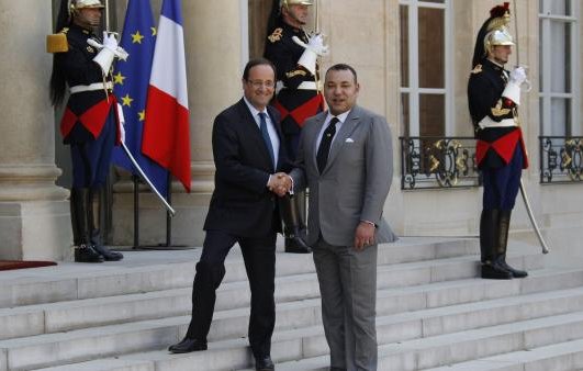 L'image montre une scène officielle à l'extérieur d'un bâtiment gouvernemental. Deux hommes, en costume, se serrent la main sur des marches, symbolisant une rencontre diplomatique. À l'arrière-plan, on peut voir des drapeaux de la France et du Maroc, ainsi que des soldats en tenue d'apparat. L'atmosphère est formelle et solennelle, reflétant l'importance de l'événement.