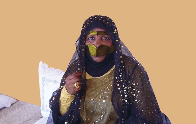 L'image montre une femme vêtue d'une tenue traditionnelle ornée de motifs et de broderies. Elle porte un voile noir qui couvre son visage en grande partie, laissant seulement ses yeux visibles, ce qui ajoute une touche de mystère. Sa robe est richement décorée, avec des couleurs sombres et des embellissements scintillants. À côté d'elle, on aperçoit des objets traditionnels, ce qui suggère une ambiance culturelle. L'arrière-plan est d'une couleur unie, mettant en valeur sa présence.