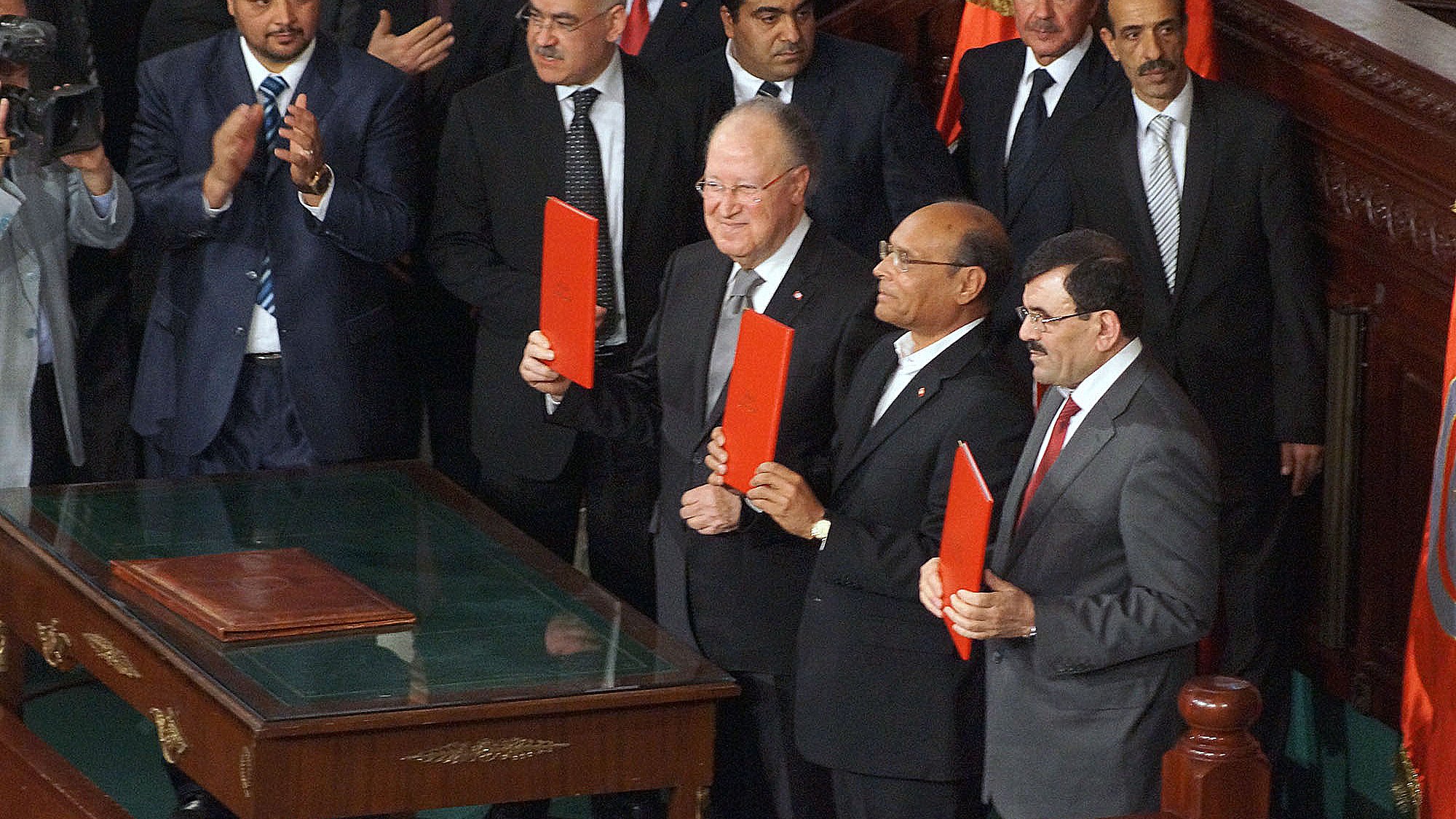 L'image montre un groupe d'hommes se tenant debout autour d'une table, tous souriants et tenant des dossiers rouges. Ils semblent être dans un cadre officiel, probablement lors d'une cérémonie ou d'une réunion importante. En arrière-plan, on peut voir d'autres personnes assises et applaudissant, ainsi que des drapeaux. L'ambiance semble solennelle et festive à la fois.