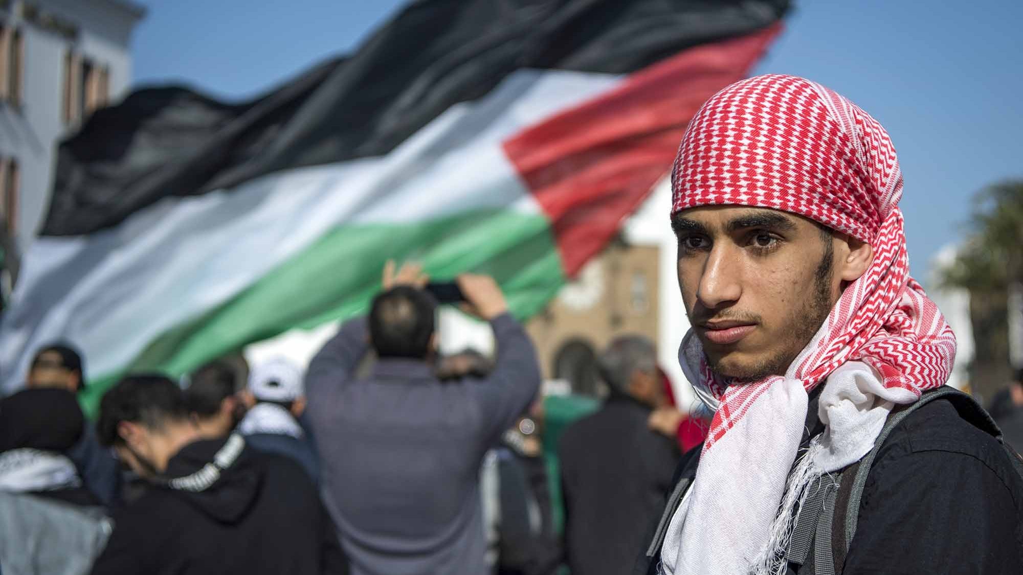 L'image montre un jeune homme portant un keffieh rouge et blanc, se tenant au premier plan. En arrière-plan, une grande foule est visible, avec un drapeau palestinien noir, blanc, vert et rouge qui flotte. L'atmosphère semble être celle d'une manifestation ou d'un rassemblement, où les participants expriment leurs convictions ou leur soutien à une cause. Le jeune homme affiche une expression sérieuse, ce qui ajoute à l'intensité de la scène.