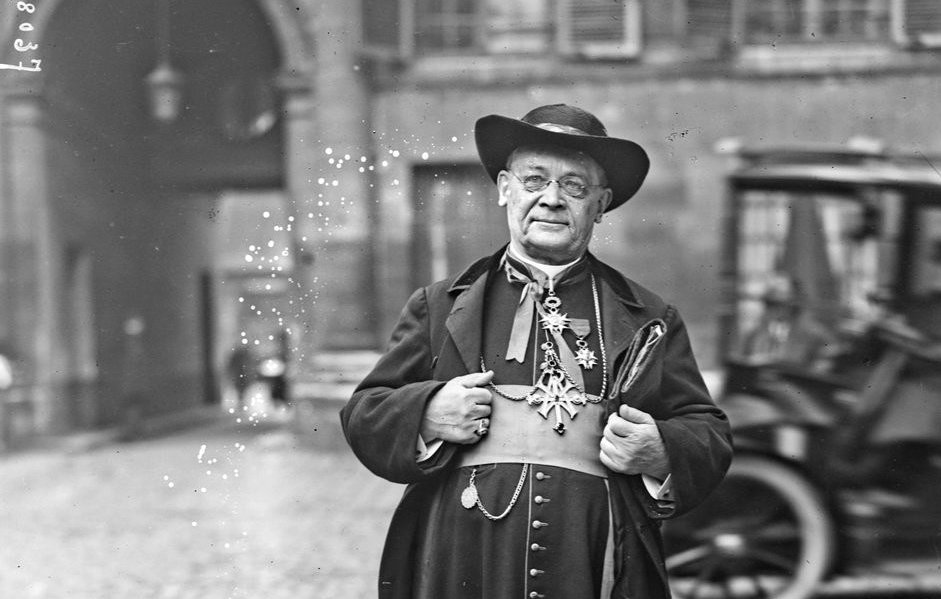 L'image montre un homme en tenue traditionnelle, probablement un ecclésiastique. Il porte une longue robe noire avec des ornements et des boutons sur le devant. Son chapeau est large et il semble poser avec une certaine dignité, les bras légèrement écartés. En arrière-plan, on aperçoit des bâtiments anciens et un véhicule à roues, ce qui suggère une ambiance historique. L'environnement de la rue est pavé, ajoutant à l'atmosphère d'époque.