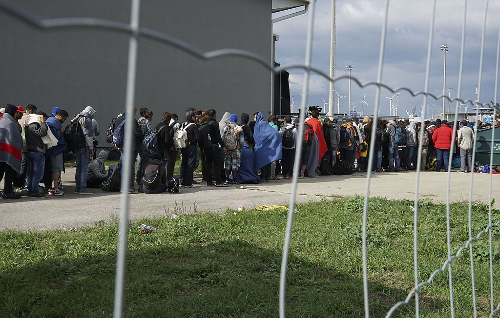 L'image montre une file de personnes faisant la queue près d'un bâtiment, probablement en attendant de recevoir de l'aide ou des services. Les personnes sont vêtues de diverses manières, avec certains portant des couvertures. On peut voir une clôture en fil de fer qui sépare le groupe du reste de l'environnement. En arrière-plan, on aperçoit des installations modernes et des éoliennes, suggérant une zone urbaine ou industrielle. Le ciel est nuageux, ce qui pourrait indiquer une atmosphère morose.
