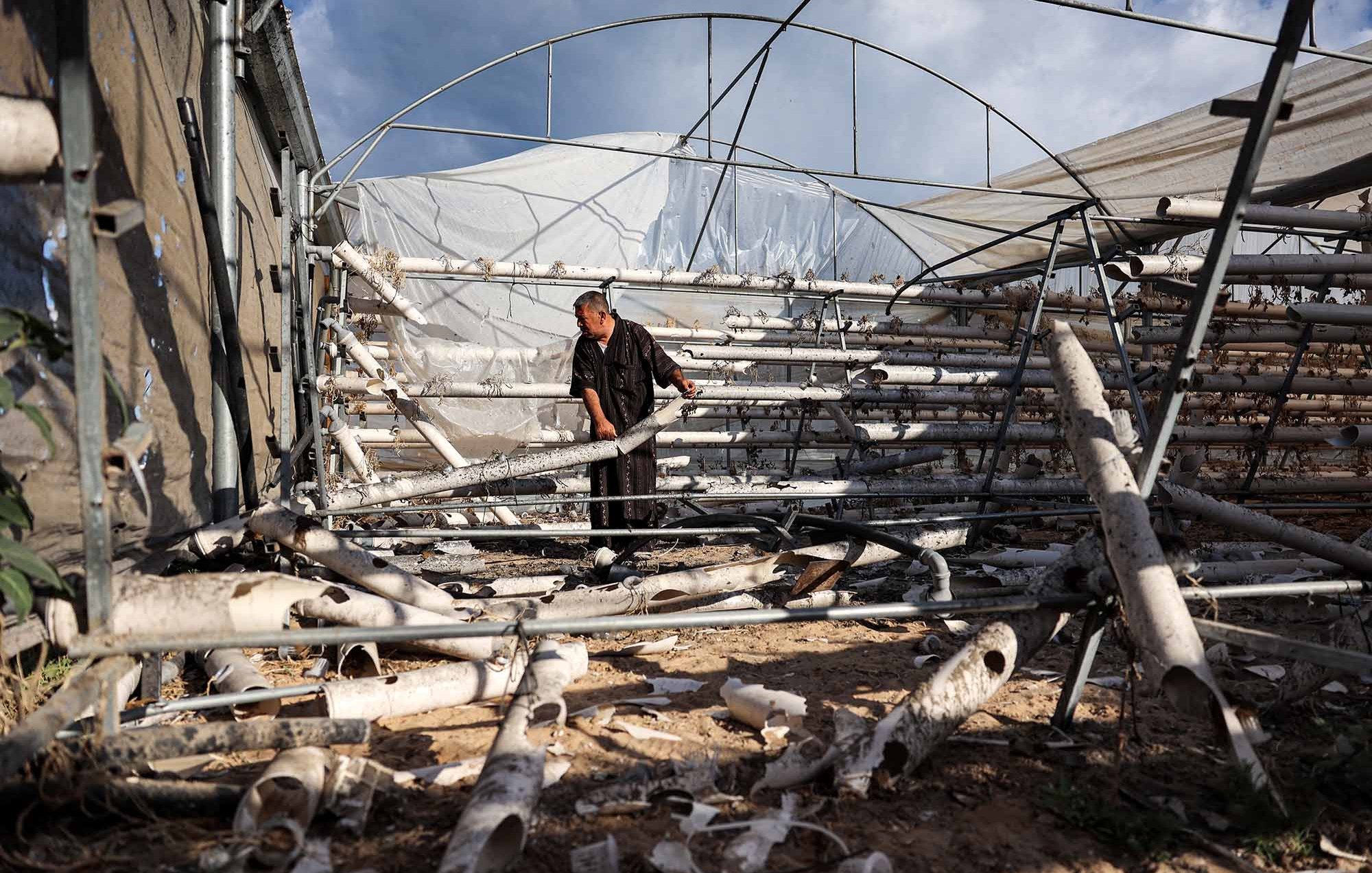 L'image montre une personne à l'intérieur d'une serre. On voit des tubes en plastique ou en métal disposés sur le sol, probablement utilisés pour des systèmes d'irrigation ou de culture. La lumière naturelle entre par les ouvertures, illuminant l'espace. L'environnement semble un peu désordonné, avec des matériaux éparpillés. Le ciel est visible à travers la structure, indiquant qu'il fait jour.