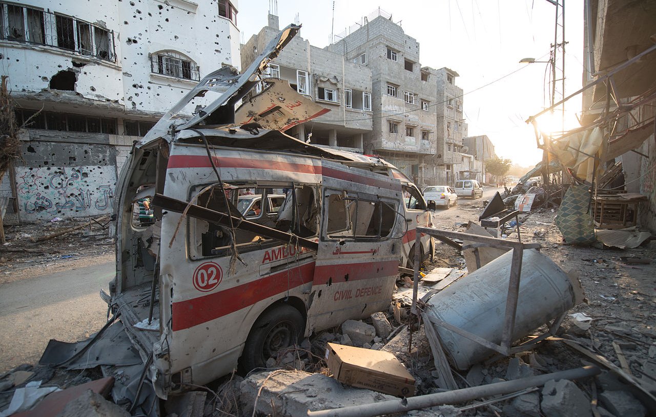 L'image montre une scène de destruction urbaine, probablement à la suite d'un conflit. On y voit une ambulance endommagée, avec sa structure déformée et son revêtement extérieur marqué par des débris. Les bâtiments environnants sont en ruine, avec des fenêtres brisées et des murs partiellement effondrés. Le sol est jonché de débris, et l'éclairage indique que c'est soit tôt le matin, soit en fin d'après-midi. L'atmosphère générale est sombre et témoigne de l'impact des violences sur la ville.