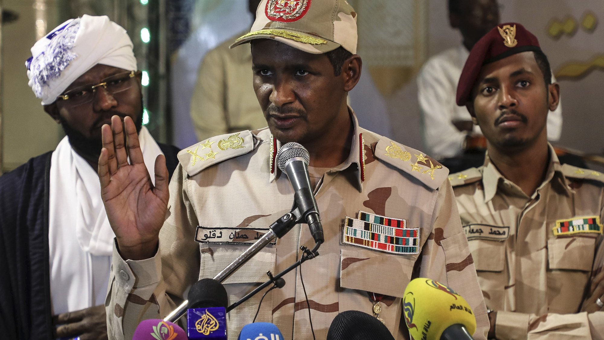 L'image montre un homme en uniforme militaire, portant une casquette décorée, qui s'exprime devant un micro lors d'une conférence de presse. Il semble être en train de faire une déclaration importante, avec une main levée. En arrière-plan, deux autres hommes sont également présents, l'un portant un chapeau traditionnel et l'autre en uniforme militaire. Divers microphones de médias sont placés devant lui. L'ambiance semble sérieuse, suggérant un contexte politique ou militaire.
