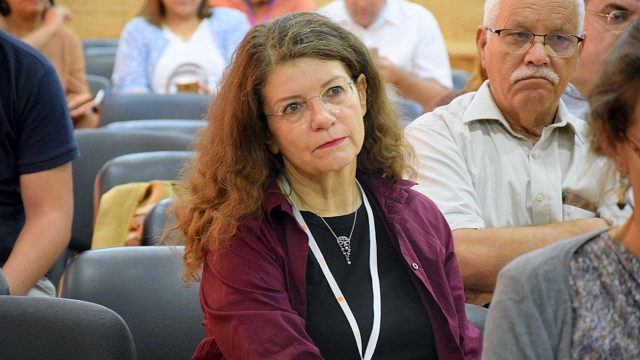 L'image montre une femme assise dans une salle de conférence ou de réunion, avec une expression pensive. Elle porte un haut de couleur sombre et a de longs cheveux bouclés. En arrière-plan, on peut voir d'autres personnes dans une atmosphère assez calme. La femme semble concentrée sur ce qui se passe autour d'elle.