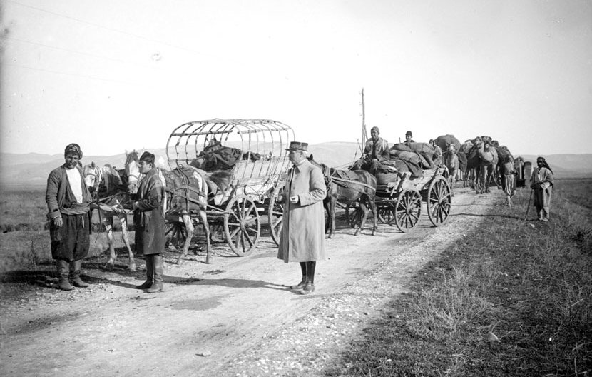 L'image montre une scène historique sur un chemin. On y voit plusieurs chariots tirés par des chevaux, ainsi que des personnes qui se déplacent. Certaines personnes sont debout près des chariots, tandis que d'autres semblent s'affairer autour des animaux. L'environnement est rural, avec des champs en arrière-plan et une atmosphère qui évoque des temps passés, probablement sur un chemin de terre. Les vêtements des personnes suggèrent une époque ancienne.