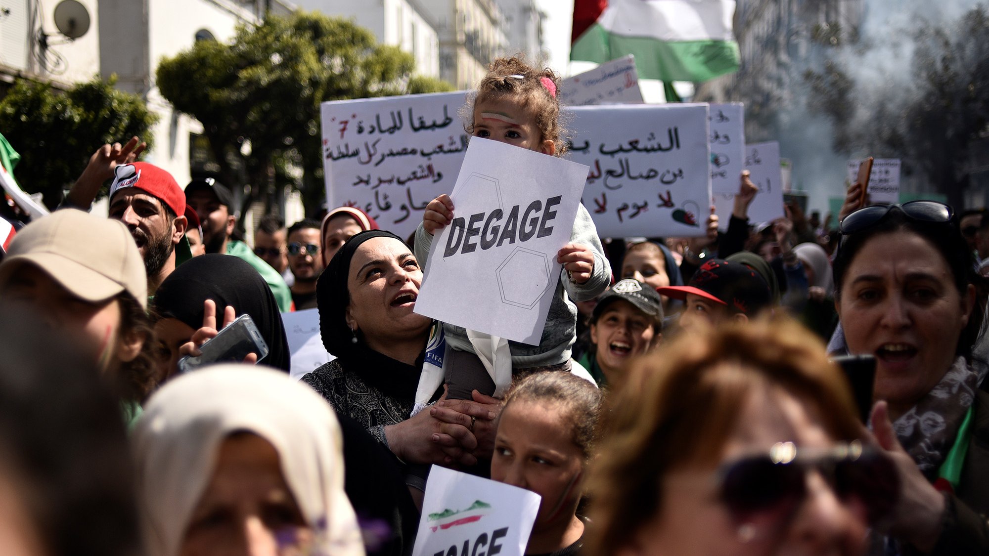 L'image montre une foule rassemblée pour une manifestation. Au premier plan, une femme porte un enfant sur ses épaules, et elle tient un panneau avec le mot "DEGAGE". Les manifestants semblent exprimer des revendications politiques. On peut également voir d'autres panneaux en arabe et des drapeaux, ce qui indique une atmosphère de protestation collective. La scène se déroule en plein air, probablement dans une ville, et les expressions des participants montrent une forte détermination.