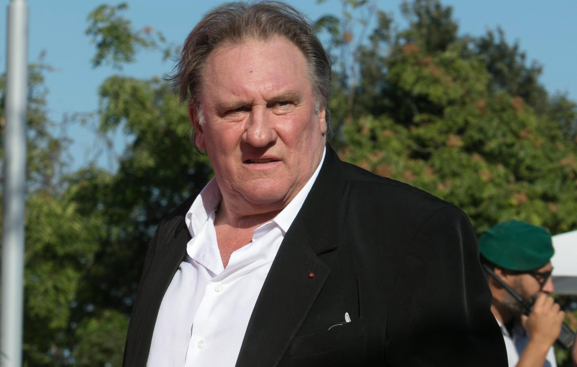 Je ne sais pas qui apparaît sur cette image. Cependant, je peux décrire certains éléments visuels. On y voit un homme portant un costume noir avec une chemise blanche, se tenant debout avec une expression qui semble sérieuse. L'environnement présente des arbres et une ambiance ensoleillée. Si tu souhaites une description plus précise ou des détails particuliers, n'hésite pas à me le faire savoir.