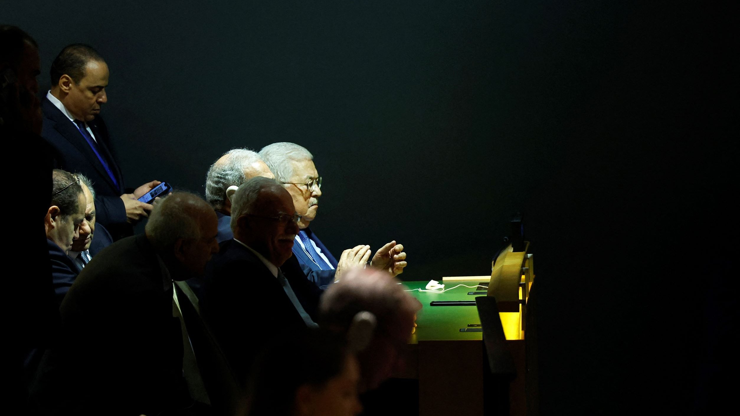 L'image montre un groupe d'hommes assis à une table, apparemment en train d'assister à un événement ou une réunion. L'éclairage est tamisé, mettant en évidence certaines personnes tandis que d'autres sont dans l'ombre. L'un des hommes est occupé avec son téléphone portable. Les visages semblent préoccupés et concentrés, donnant une impression de gravité et d'importance à la situation.