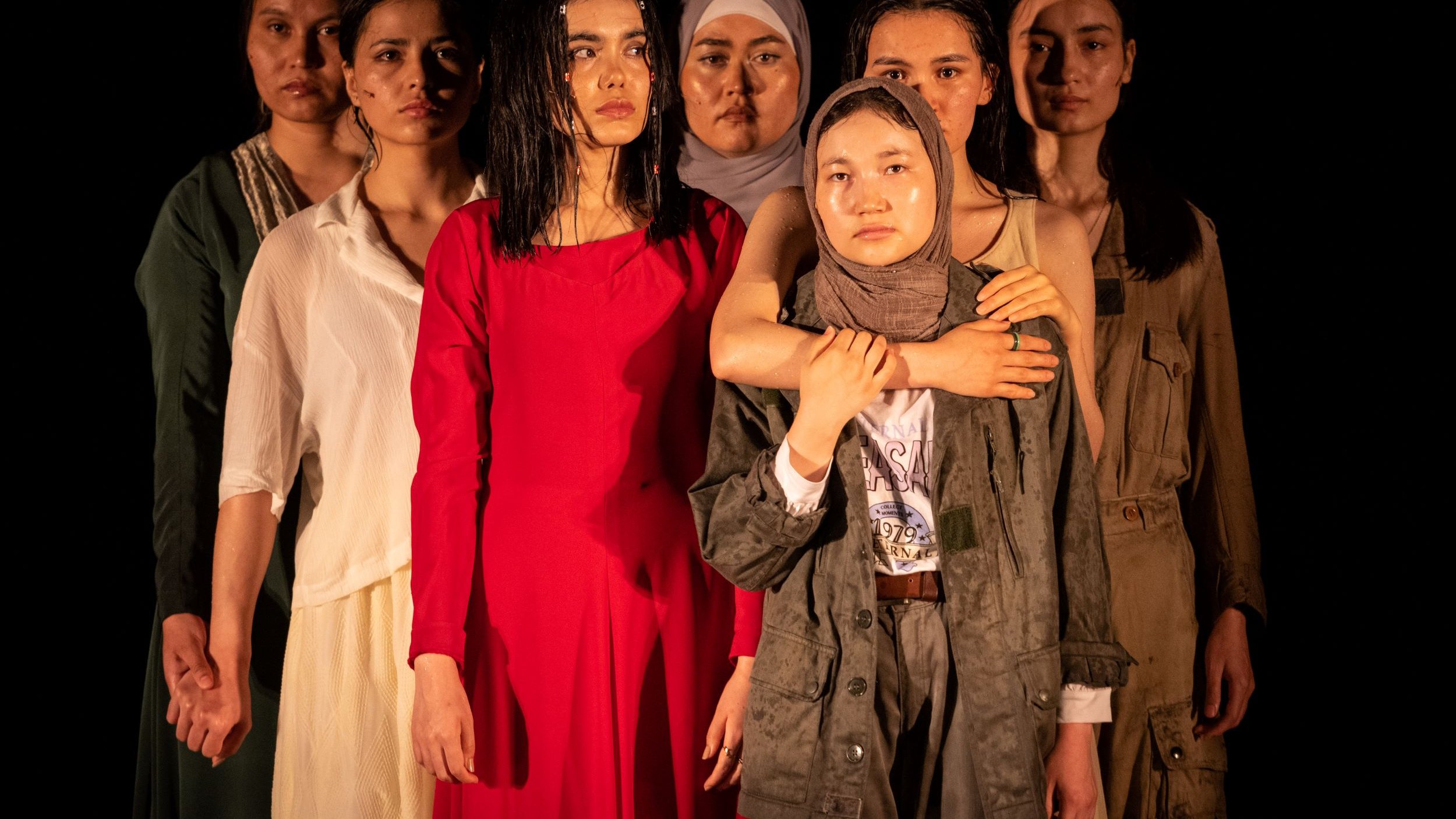 L'image montre un groupe de sept femmes, chacune portant des vêtements différents. Elles se tiennent en rang, avec des expressions sérieuses sur leurs visages. La femme au centre, vêtue d'une robe rouge, attire particulièrement l'attention. Les autres femmes, portant des vêtements variés allant de styles modernes à plus traditionnels, affichent une diversité de couleurs et de textures. L'éclairage met en valeur leurs visages et leurs postures, créant une atmosphère à la fois forte et émotive.