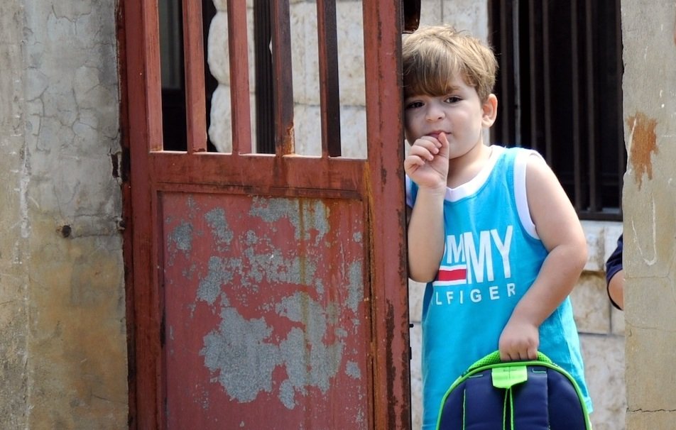 L'image montre un jeune garçon se tenant dans l'encadrement d'une porte. Il porte un débardeur bleu avec des inscriptions et tient un sac à dos coloré. Le garçon semble pensif, en train de sucer son pouce. La porte derrière lui est rustique et en métal, avec des jeux d'ombre et de lumière. L'environnement semble simple et authentique, suggérant une ambiance tranquille.
