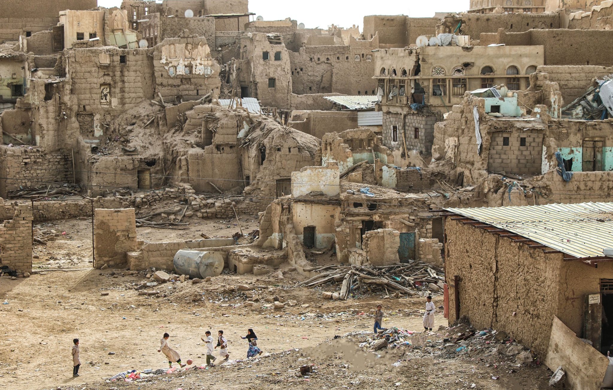 L'image montre un paysage urbain en ruines, typique d'une zone désertique. On peut voir des bâtiments en terre et en briques, avec des murs partiellement effondrés. Les structures semblent abandonnées et en mauvais état, certaines présentant des ouvertures et des décombres. Au premier plan, plusieurs enfants jouent, ce qui apporte une touche de vie à ce décor désertique. Le sol est jonché de déchets, et le ciel est clair, ce qui accentue l'aspect aride de l'environnement.