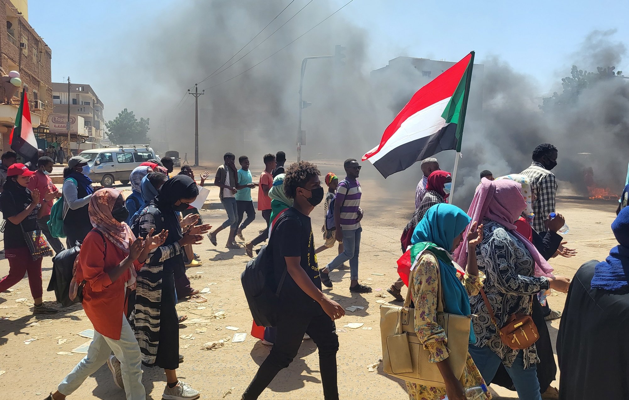 Manifestation dans la rue, fumée, drapeau soudanais, personnes marchant ensemble.