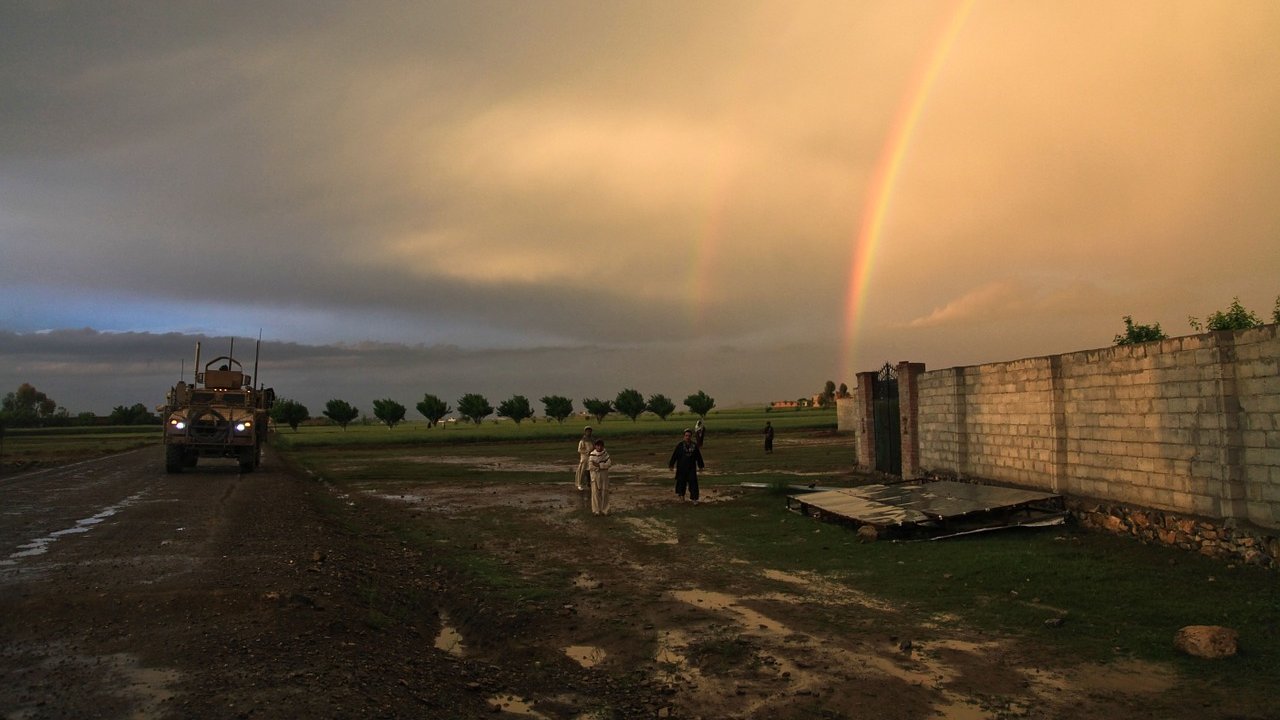 L'image montre un paysage rural sous un ciel dramatique, avec des nuages sombres et un ciel éclairci. On peut voir un arc-en-ciel double qui s'étend dans le ciel, ajoutant une touche de couleur. À gauche, un véhicule militaire est garé sur un chemin boueux, tandis qu'à droite, deux personnes marchent près d'un mur en béton. Le sol est humide, témoignant d'une récente pluie, et des arbres sont visibles en arrière-plan, soulignant une atmosphère paisible malgré la présence militaire.