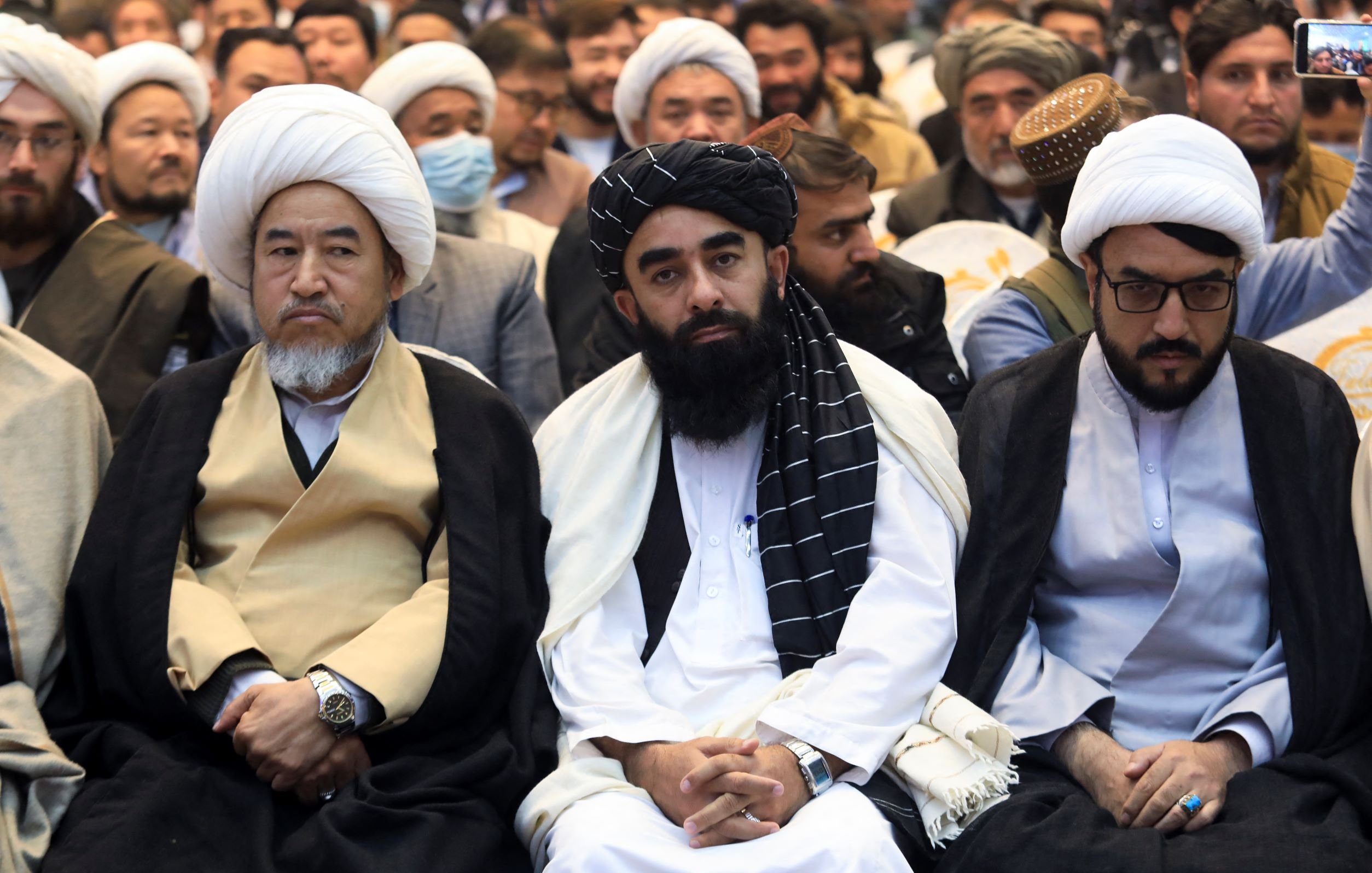 L'image montre un groupe de personnes assises, portant des vêtements traditionnels. Ils semblent participer à un événement ou une réunion. Certains d'entre eux portent des turbans ou des couvre-chefs, et l'ambiance semble solennelle. Les visages expriment diverses émotions, allant d'une concentration sérieuse à une attitude réfléchie. L'arrière-plan indique qu'il y a d'autres personnes présentes dans l'événement.